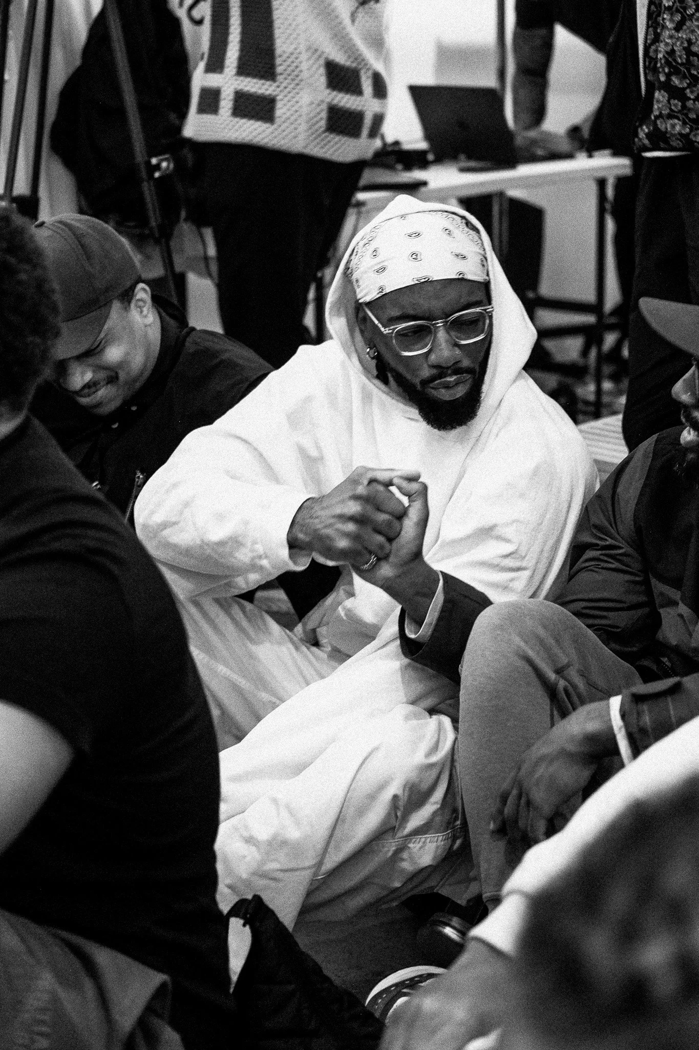 Two men handshake at a crowded indoor event; one man wears glasses, a headscarf, and a white hooded garment, while the other wears a dark cap and jacket.