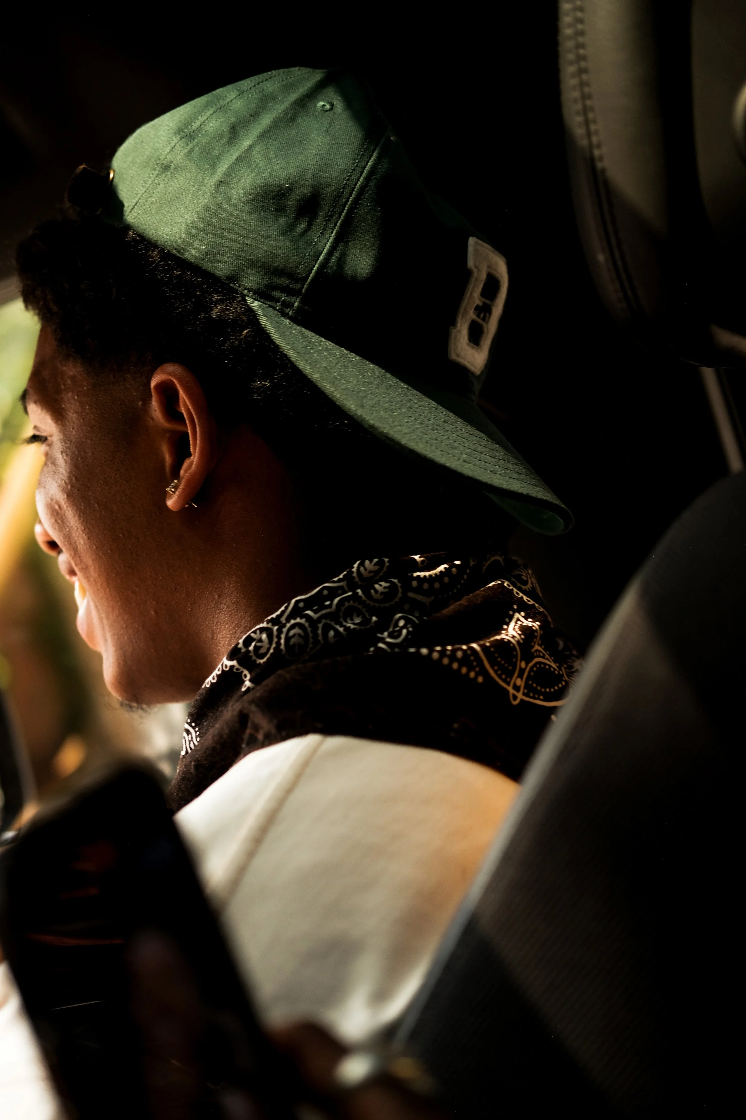 A person with a green baseball cap, earrings, and a bandana, smiling and looking at a phone, sitting in a vehicle.