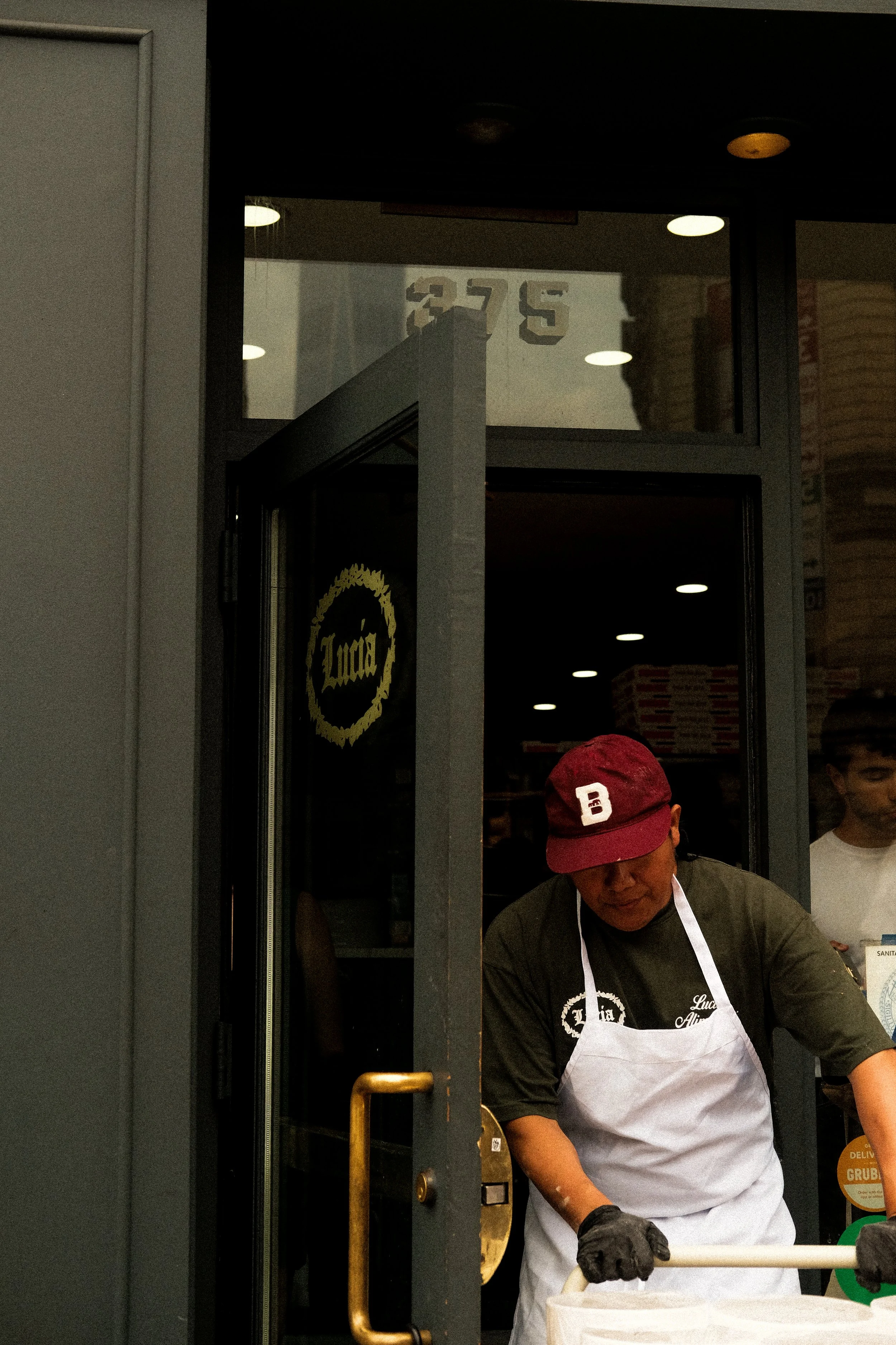 A man wearing a red cap, glasses, a dark shirt, black gloves, and a white apron operates a pizza-making station at a restaurant. The restaurant's window has the name 'Luca's' on it.