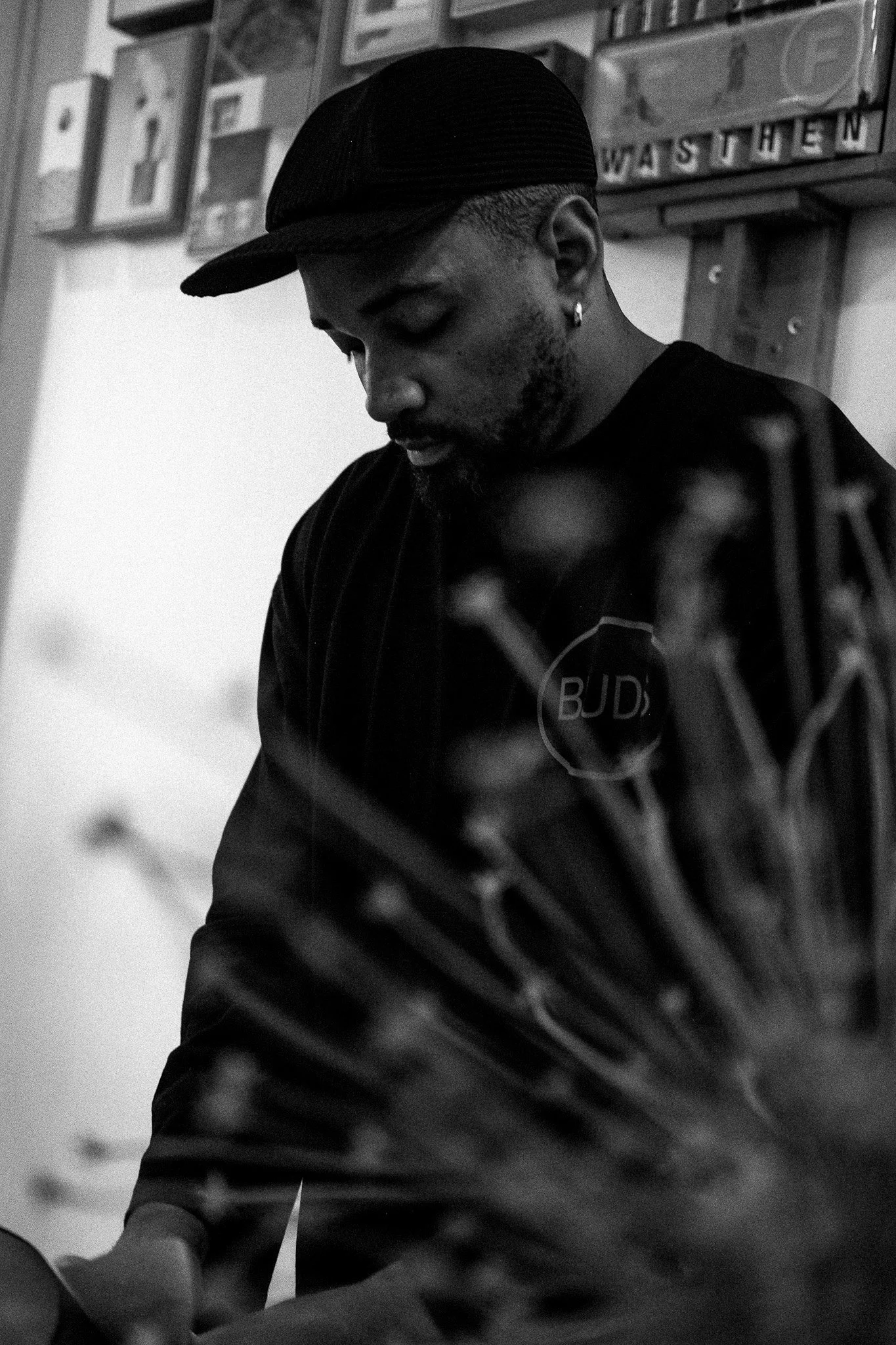 A young man with a beard wearing a black cap and black shirt looking down in black and white. In the foreground, out of focus, are the tips of a firework in a fireworks display.