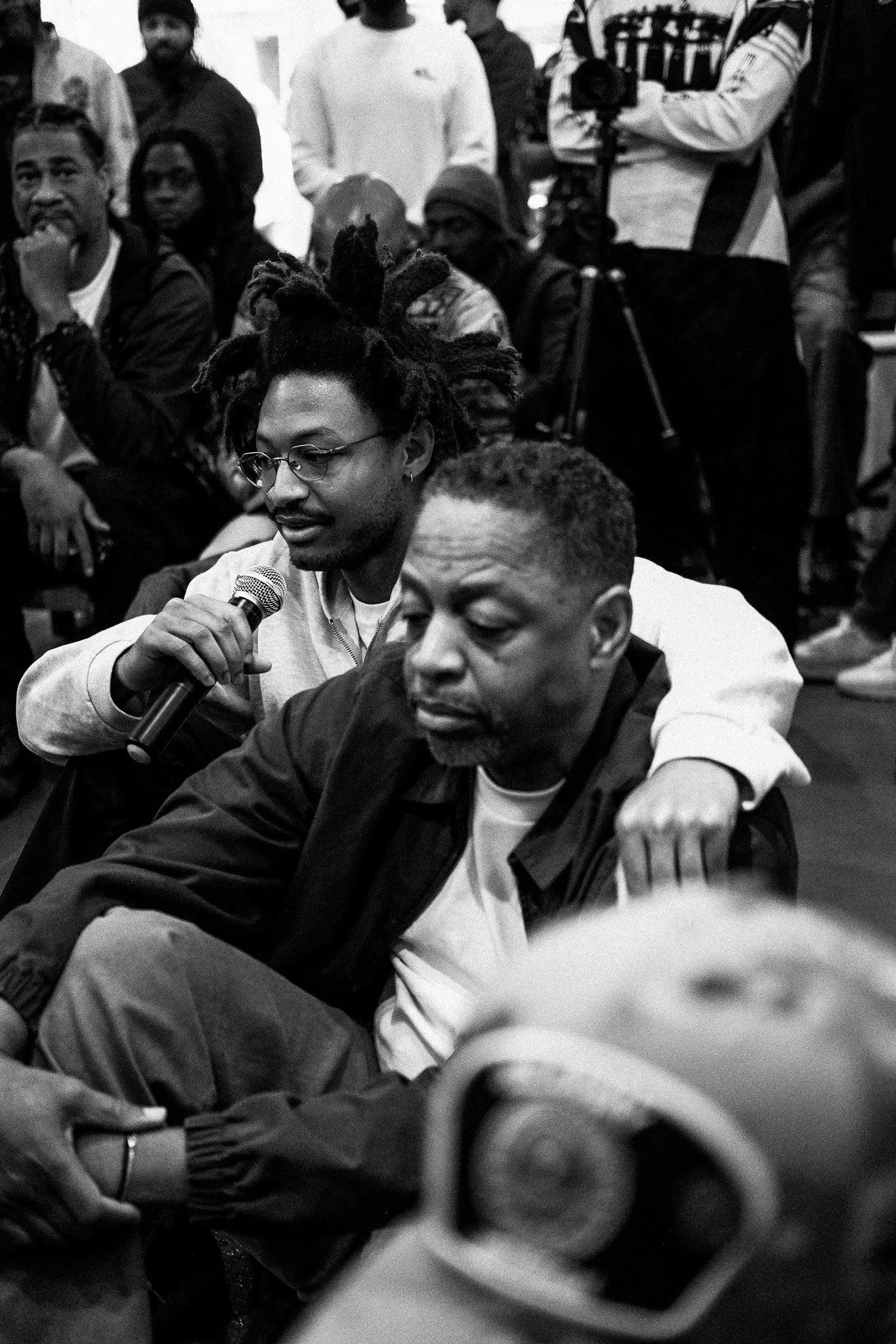 Two men sit closely together, with the man on the left holding a microphone and resting his arm on the other man's shoulders. Several people are seated and standing in the background, some observing the scene.