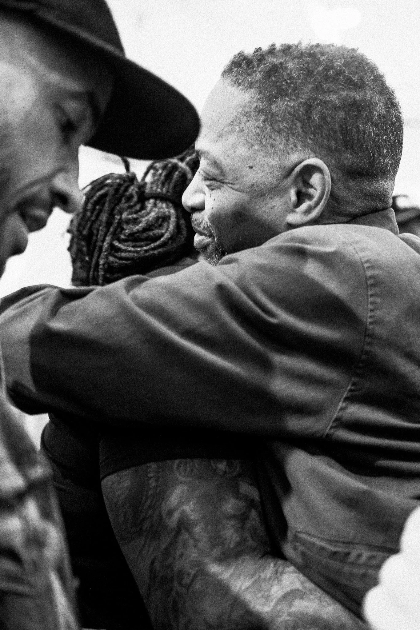 Two men hugging and smiling, one with a beard and the other wearing a hat, in a close and friendly embrace.