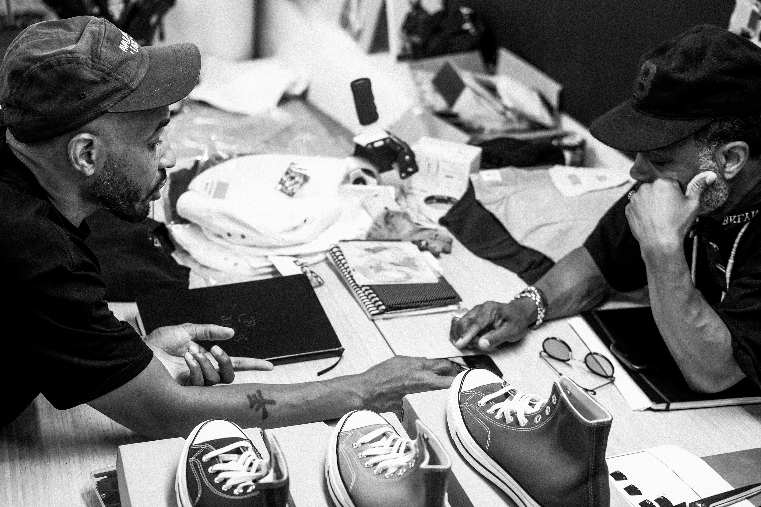 Two men engaged in a serious conversation at a cluttered table, with sneakers, glasses, notebooks, and various objects in the background.