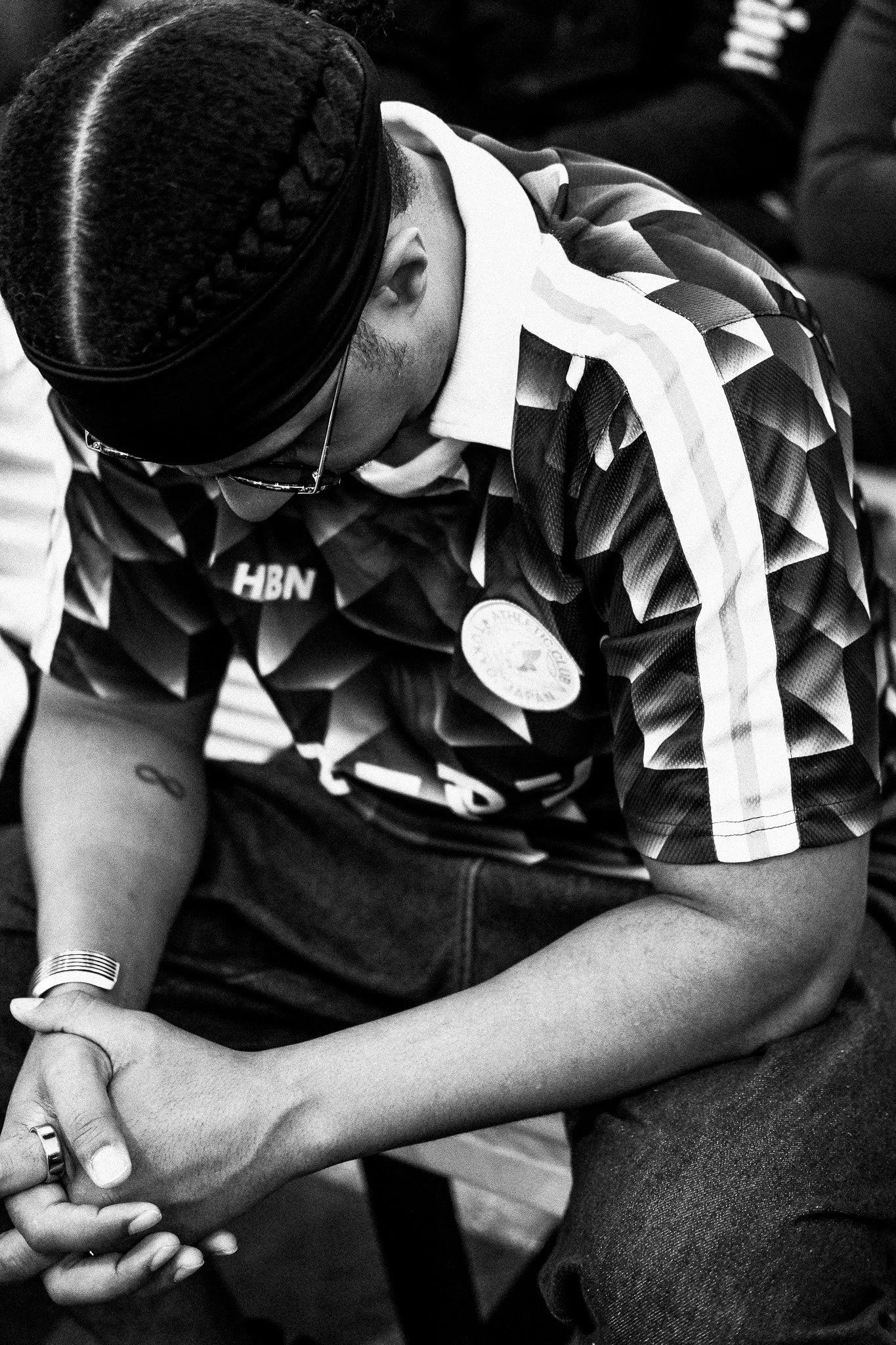 A person with braided hair, wearing glasses, a patterned sports jersey, and jeans, sitting with their hands clasped together, looking downward.