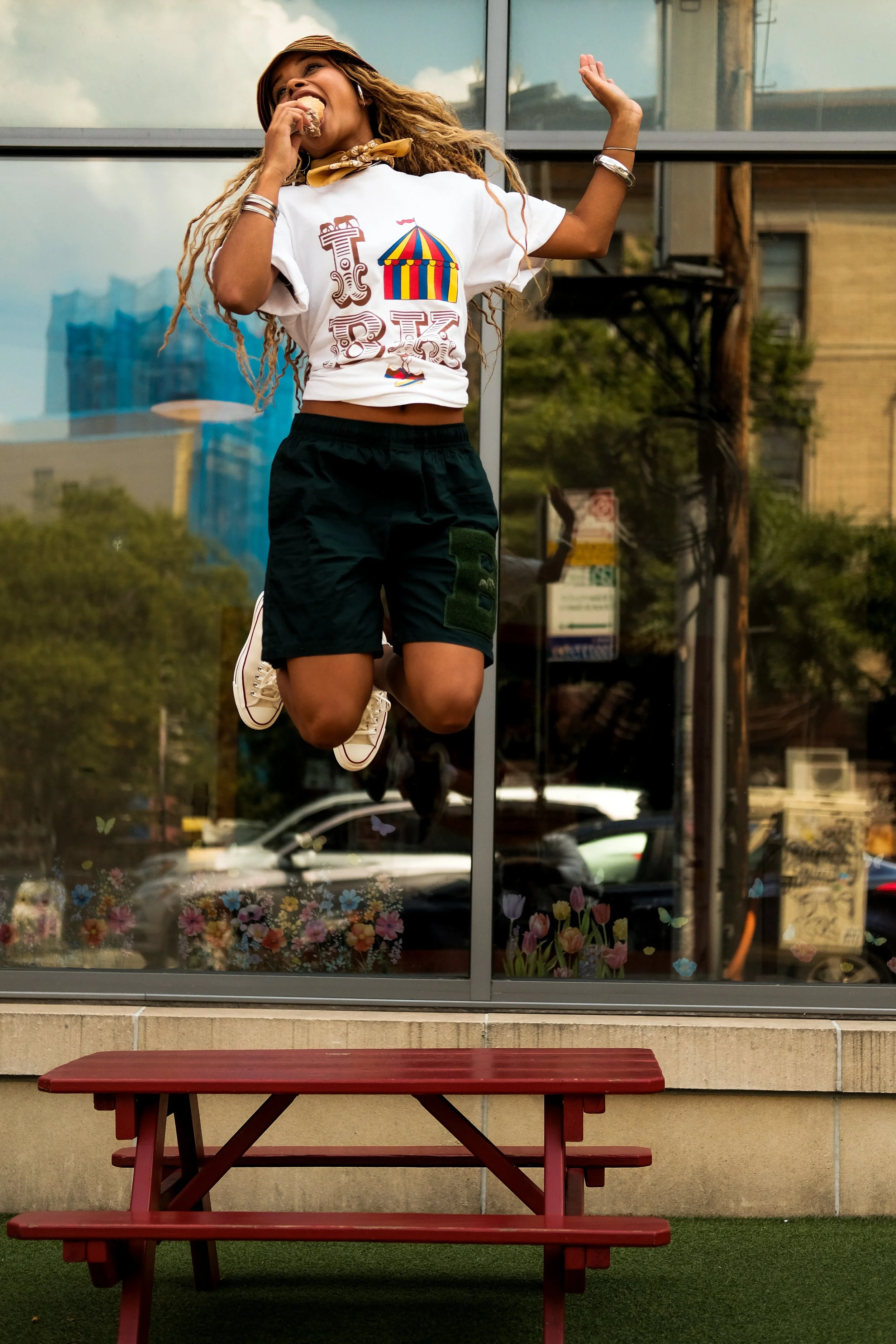 A young woman with long curly hair, wearing a hat, a white graphic t-shirt, black shorts, and sneakers, is jumping in mid-air outside a glass window, with a red picnic table in the foreground and buildings, trees, and cars reflected in the background