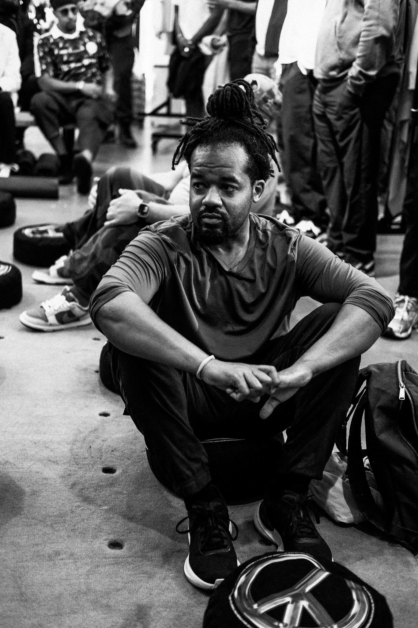 A man with dreadlocks sitting on the floor, surrounded by people in a casual setting, possibly an indoor event or gathering.