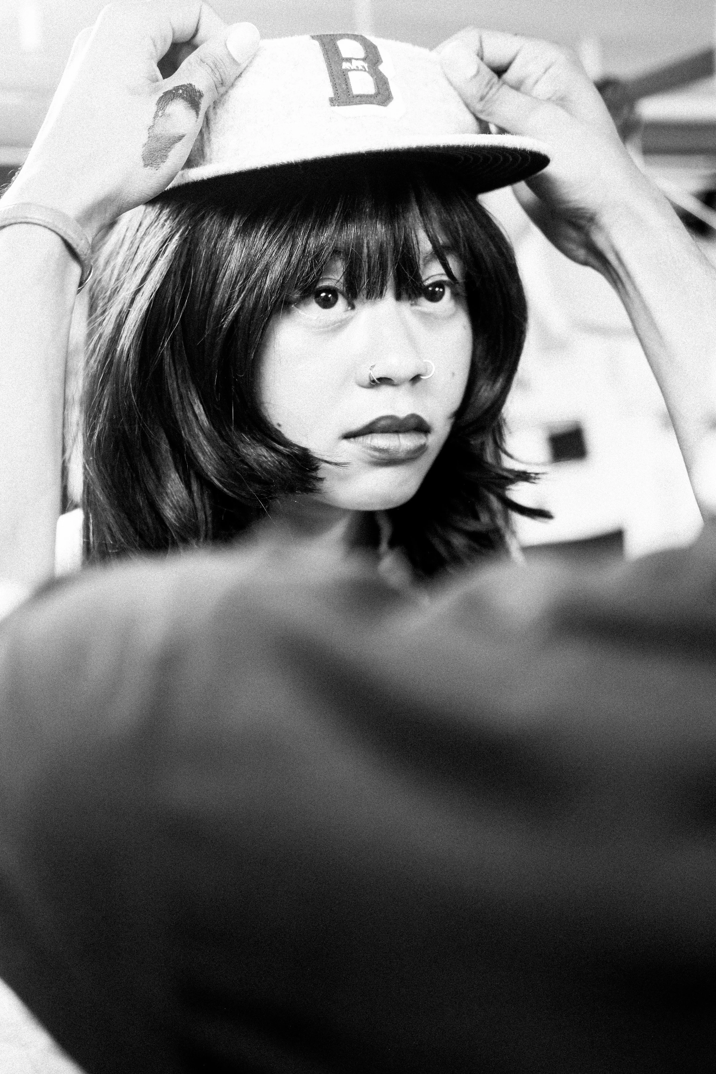 A young woman with shoulder-length dark hair and nose rings is having a baseball cap placed on her head by someone out of frame. The scene is in black and white.