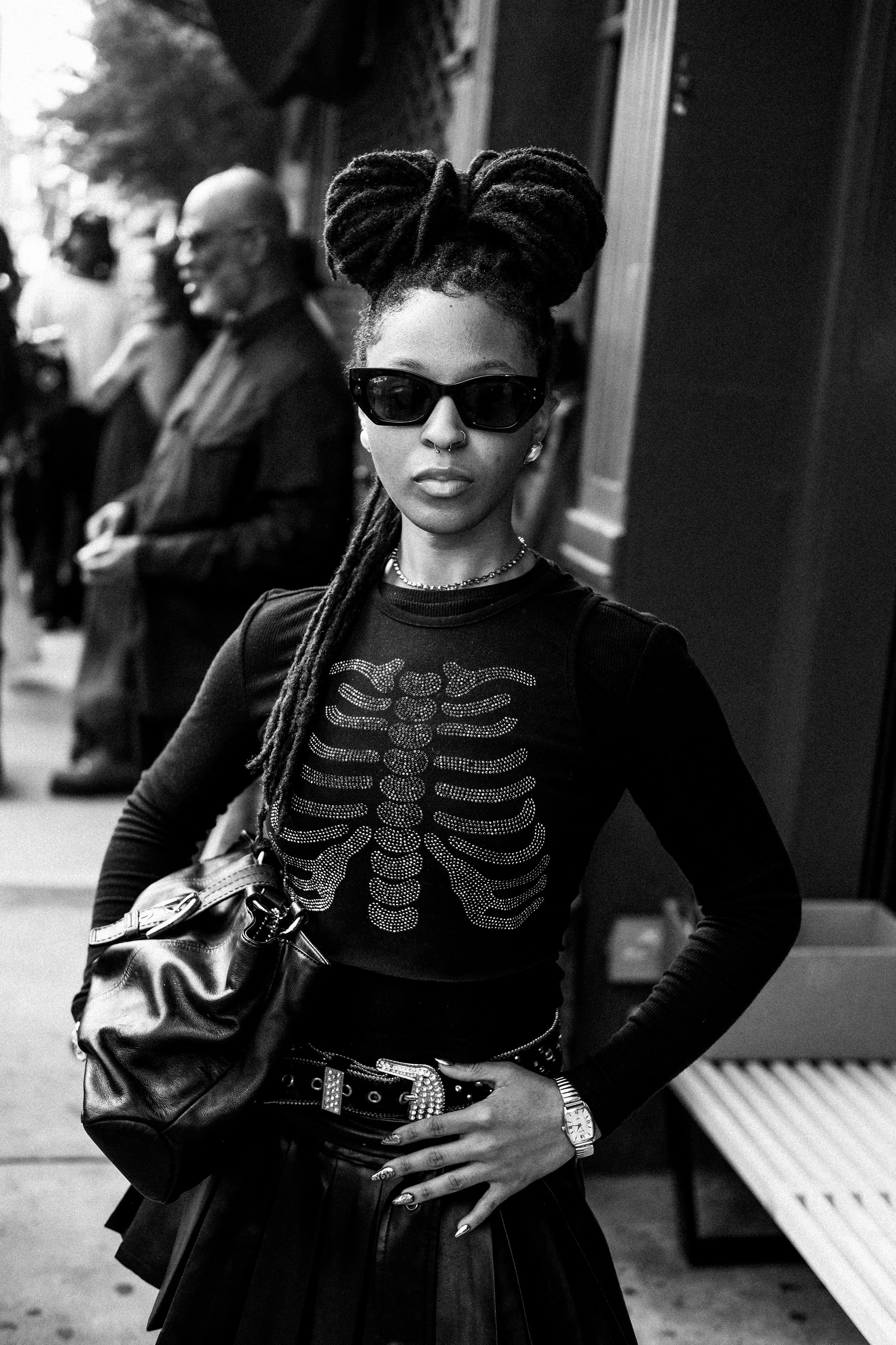 A woman with dreadlocks styled in an updo, wearing sunglasses, a nose ring, and a black long-sleeve shirt with a rhinestone skeleton design, standing on a city sidewalk with people in the background.