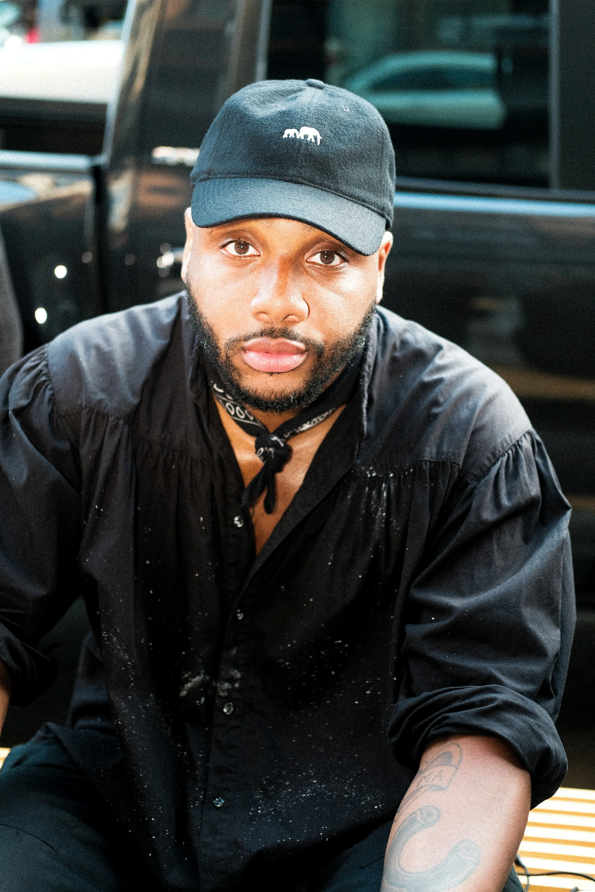 A young man with a beard wearing a black baseball cap and black shirt, sitting outdoors in front of a black vehicle.