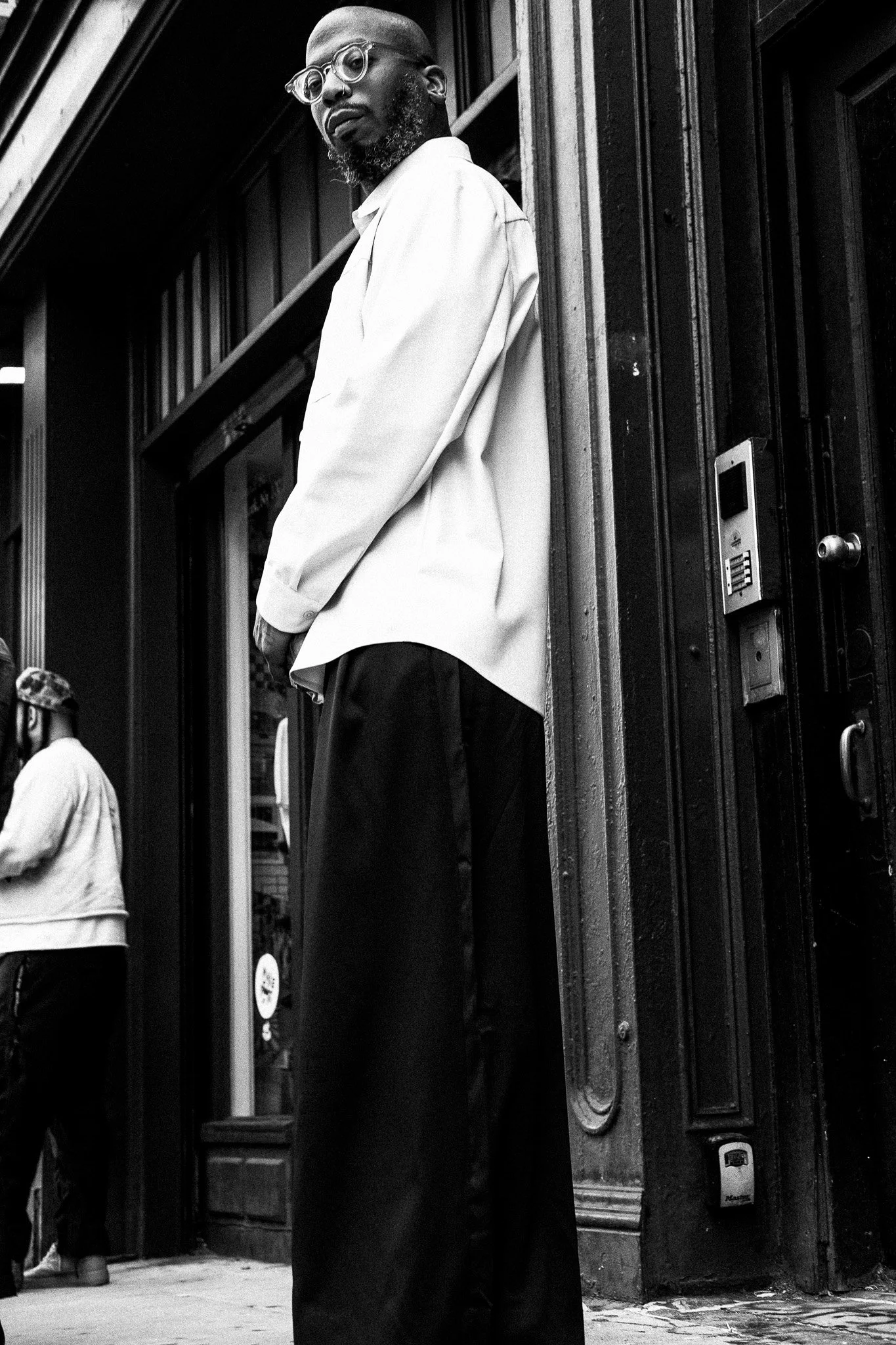 A man with glasses and a beard standing on a city street corner in black-and-white photograph.