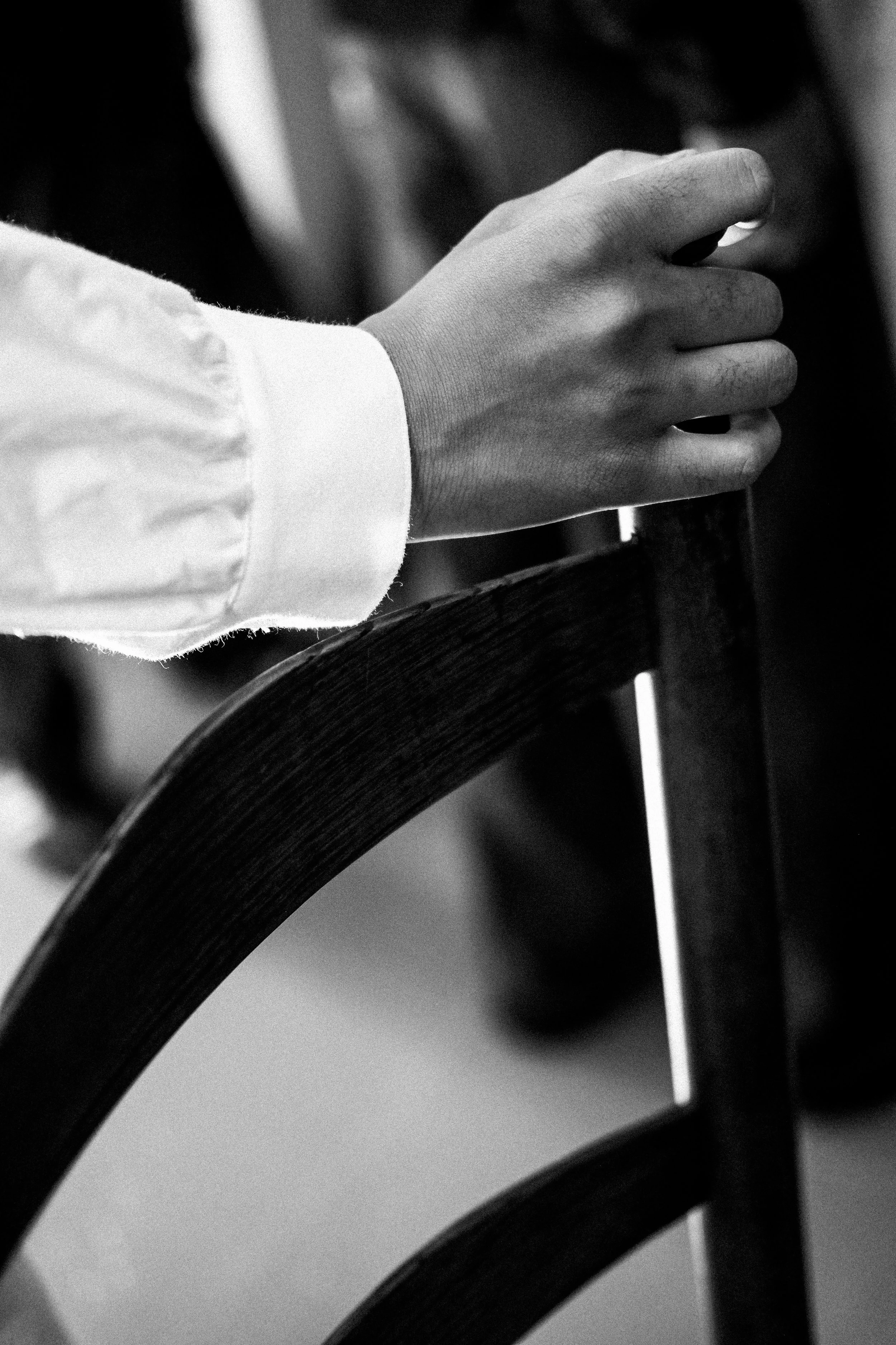 A person's hand resting on a wooden chair back, wearing a long-sleeved white shirt, in black and white.