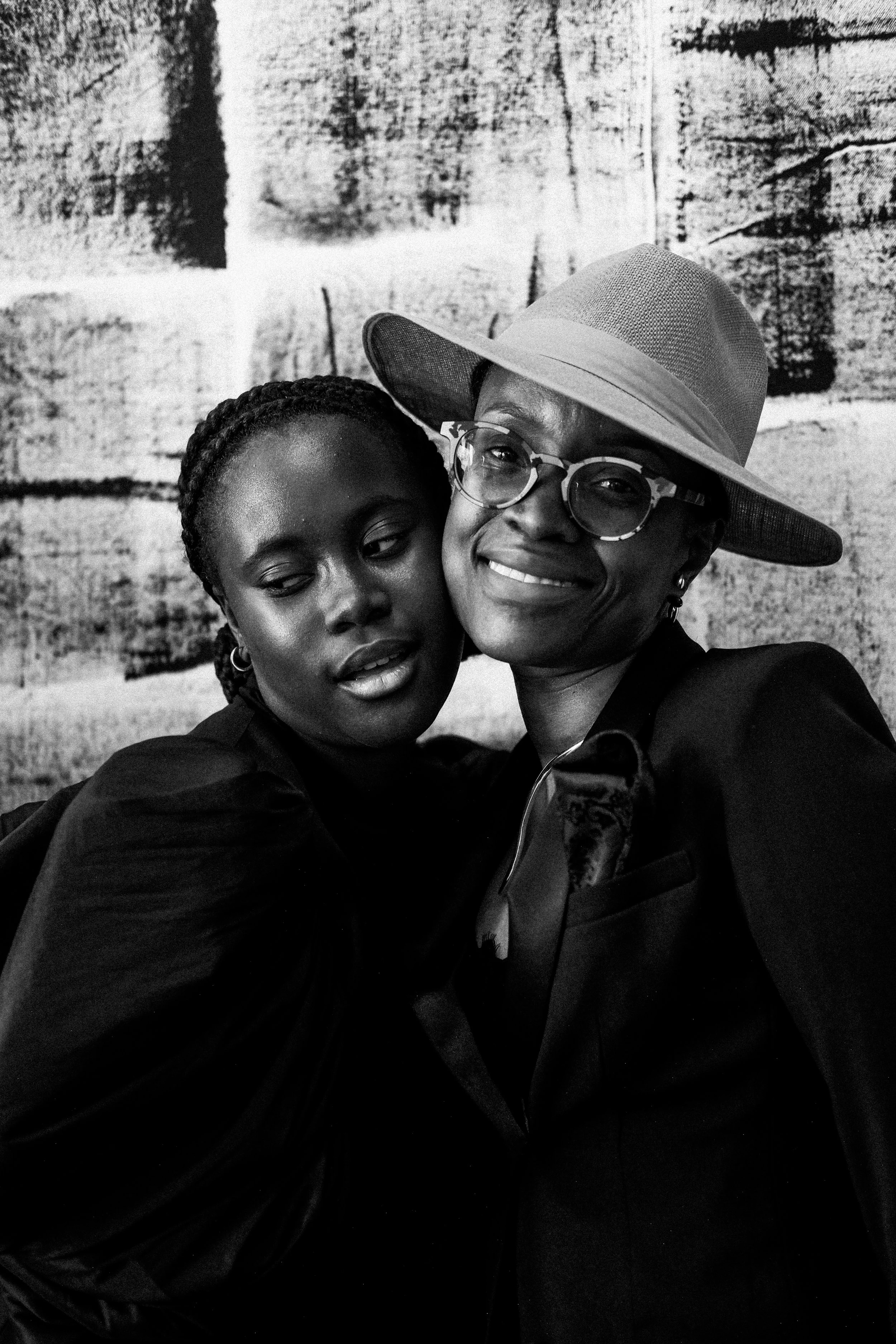 Two women embracing, one wearing a wide-brim hat and glasses, the other with braided hair, both in dark clothing, against a textured background.