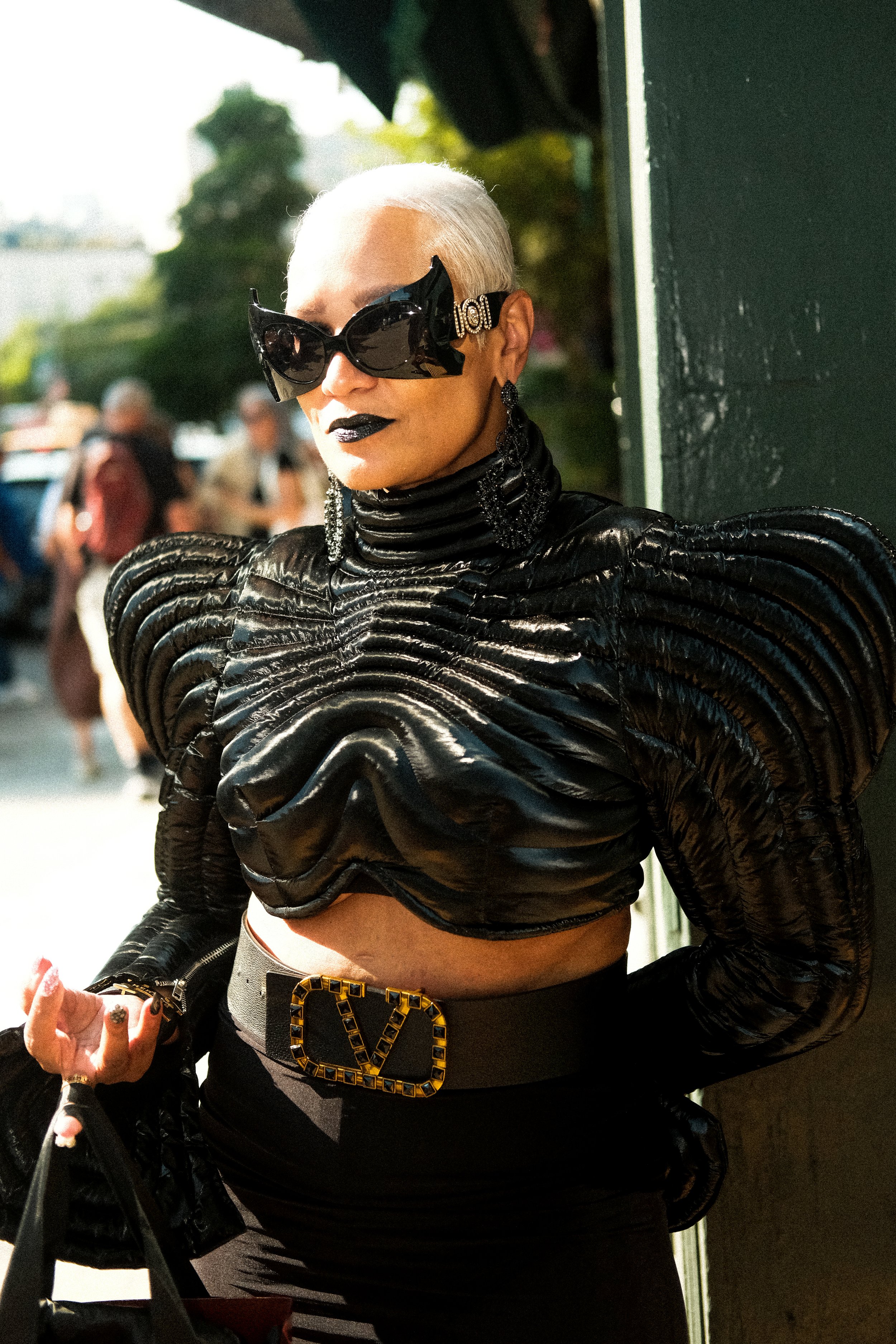 A woman with platinum blonde hair, wearing black sunglasses, black lipstick, and statement earrings, dressed in a textured black leather top with exaggerated shoulders and a belt with a gold logo.