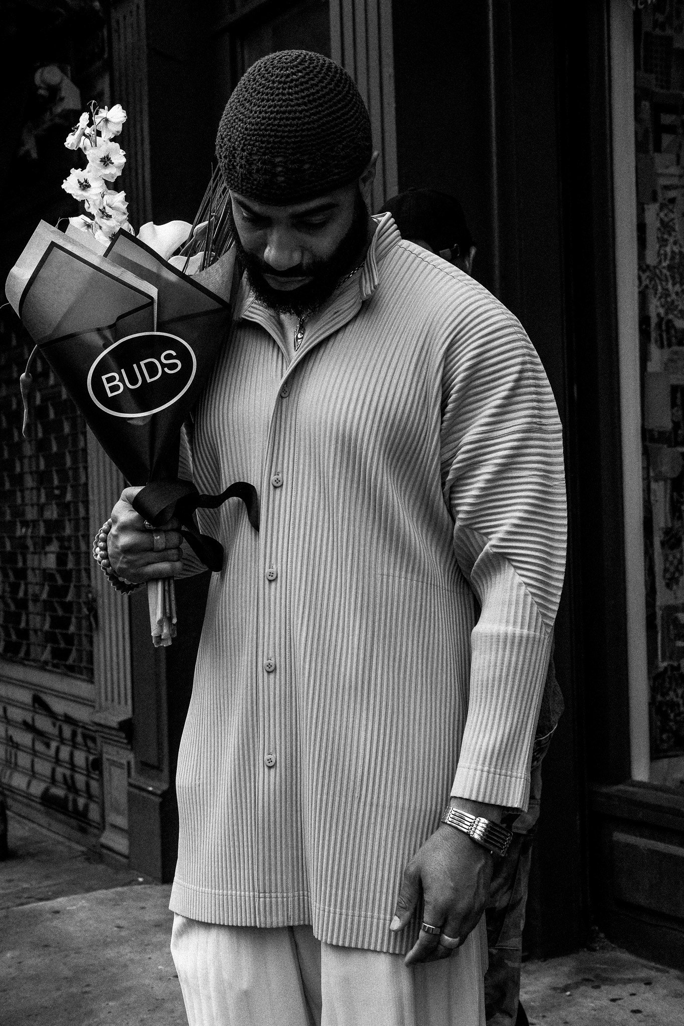 A man in a striped shirt holding a bouquet of flowers marked 'BUDS' on a city sidewalk.