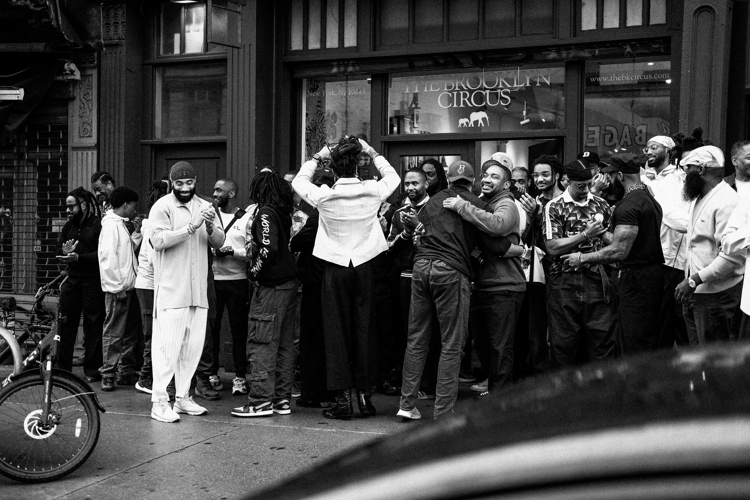 A group of people gathered outside a building with a sign that reads 'The Brooklyn Circus'. Some are smiling and interacting with each other, creating a lively, joyful scene on the street.
