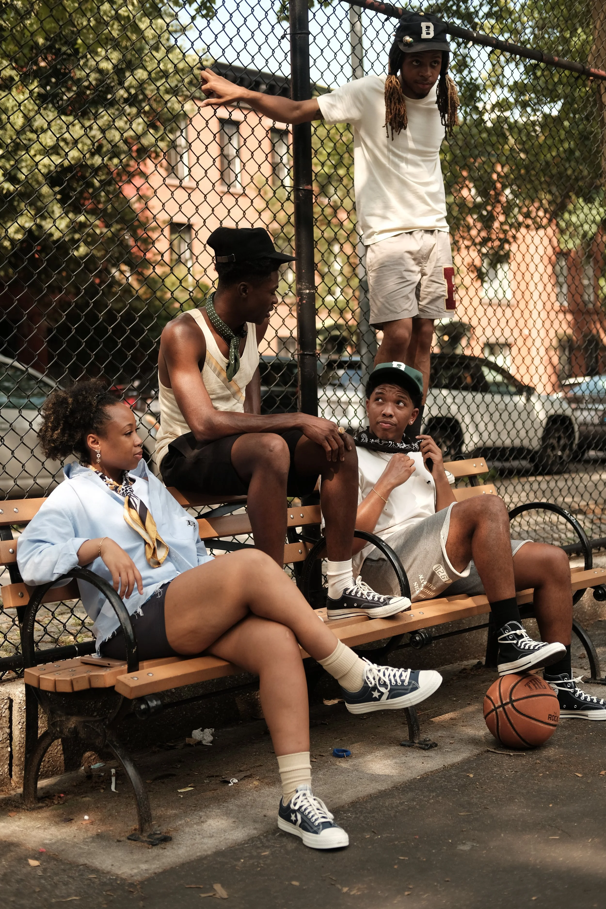 Four young friends hanging out on a park bench near a chain-link fence. One standing on the bench, while two sit on the bench, and one young woman sits at the edge with crossed legs, holding a basketball.