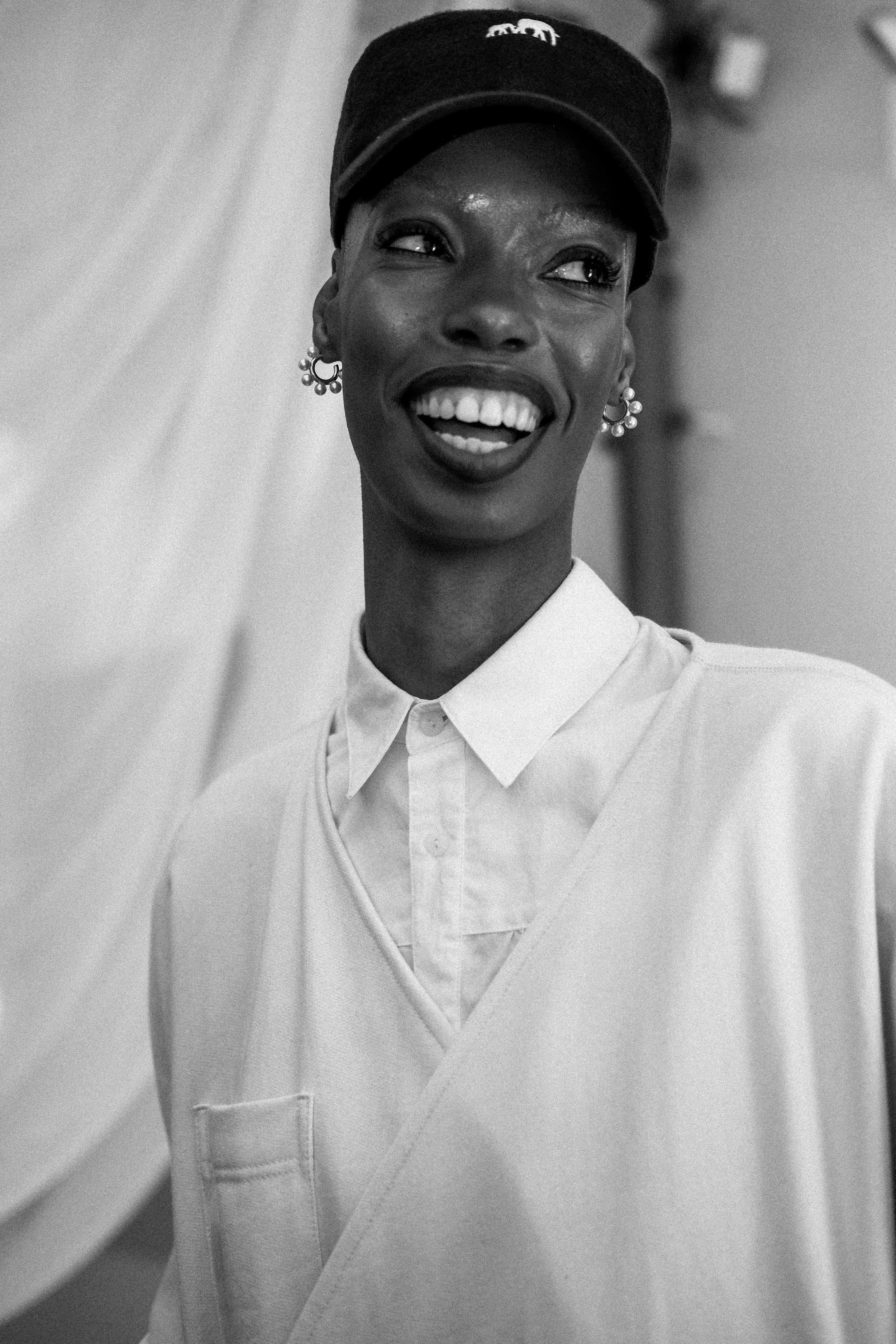 Black and white photo of a smiling woman wearing a baseball cap, earrings, a collared shirt, and a large coat or apron.