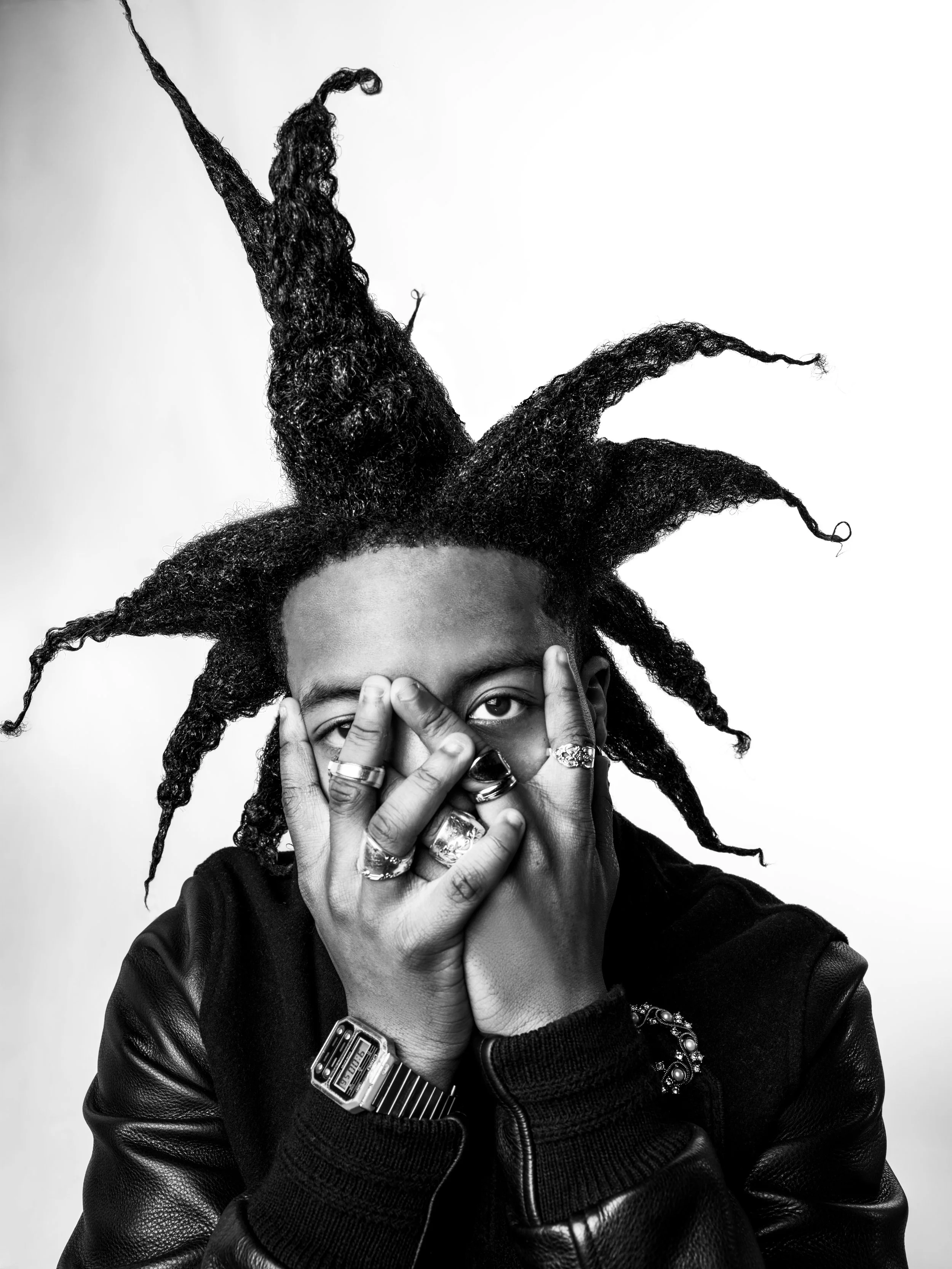 A person with dreadlocked hair, wearing multiple rings and a leather jacket, covers part of their face with their hands and looks at the camera.