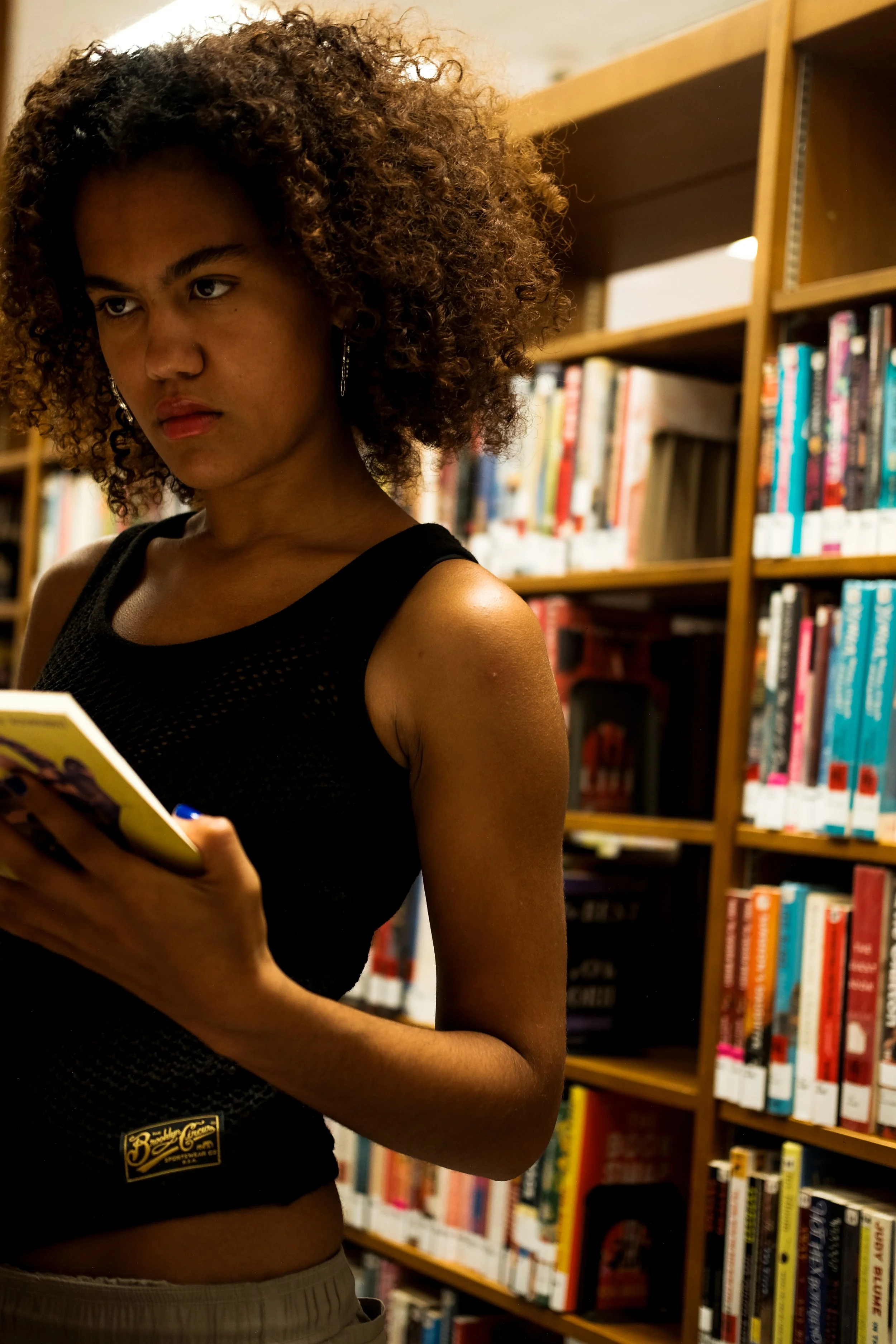 A young woman with curly hair, wearing a black sleeveless top, appears to be reading a book in a library.