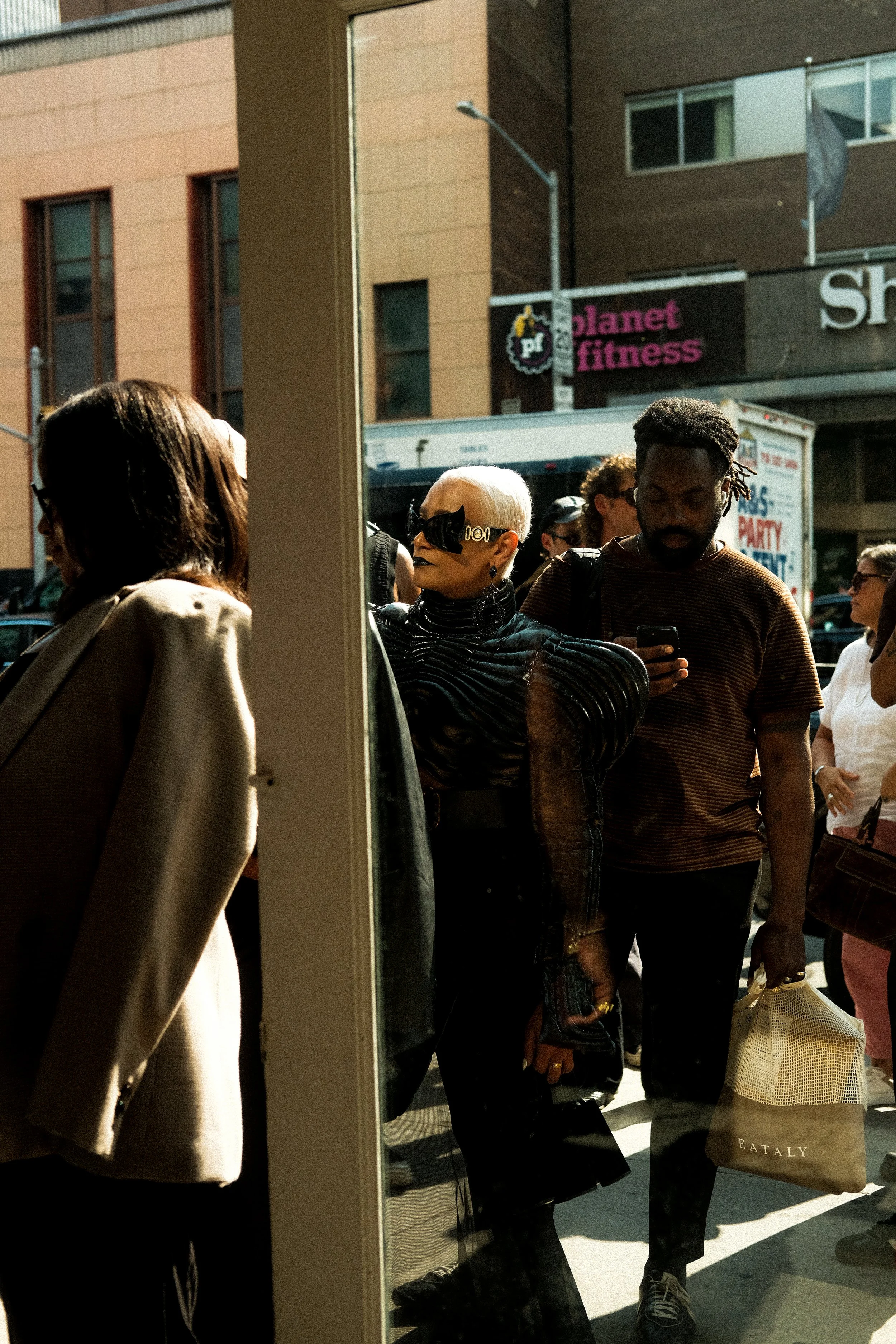 People waiting outdoors, seen through a mirror, on a city sidewalk with storefronts in the background.