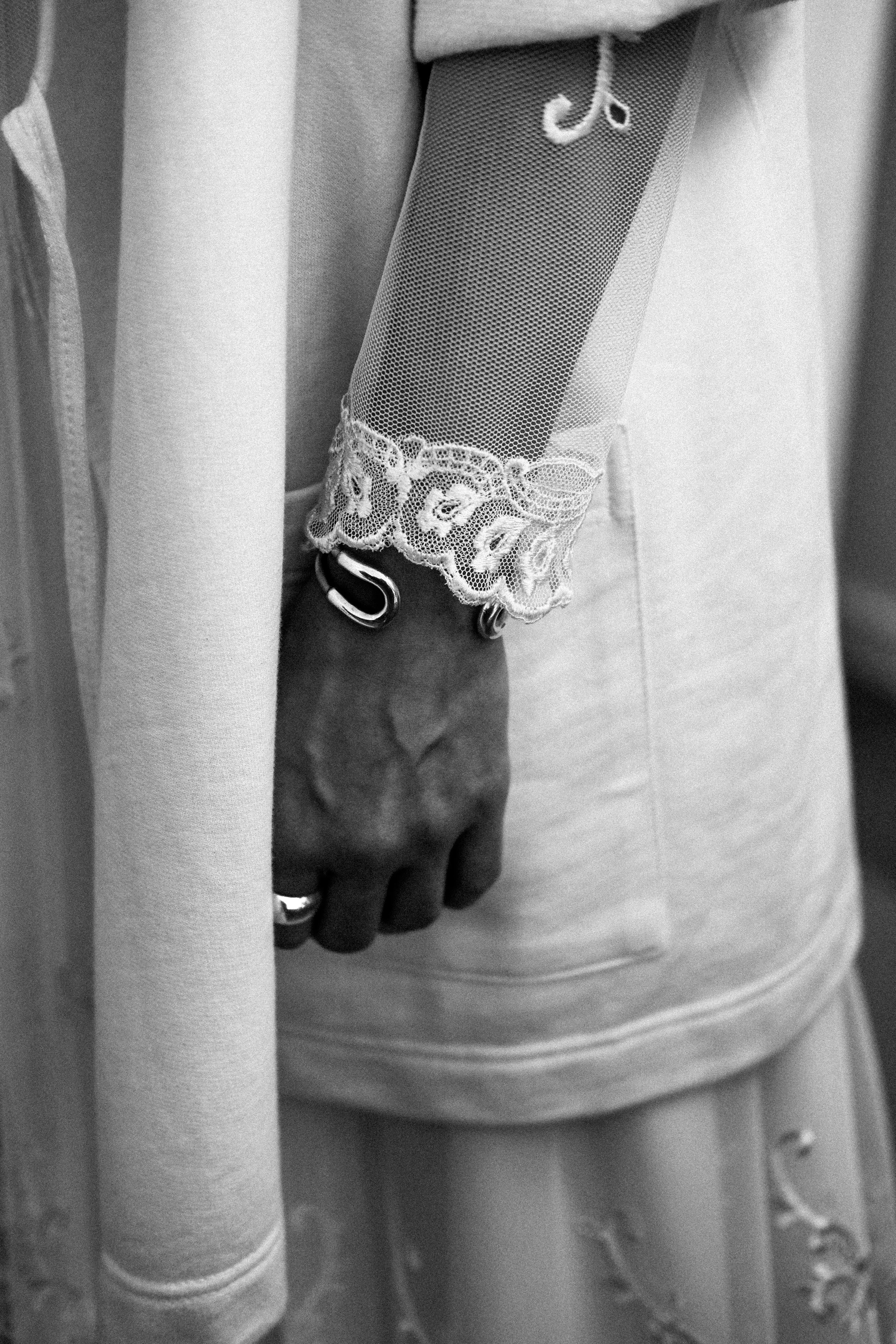 Close-up of a person's hand resting against their side, showcasing a decorative lace cuff on their sleeve and metallic safety pins on their wrist.