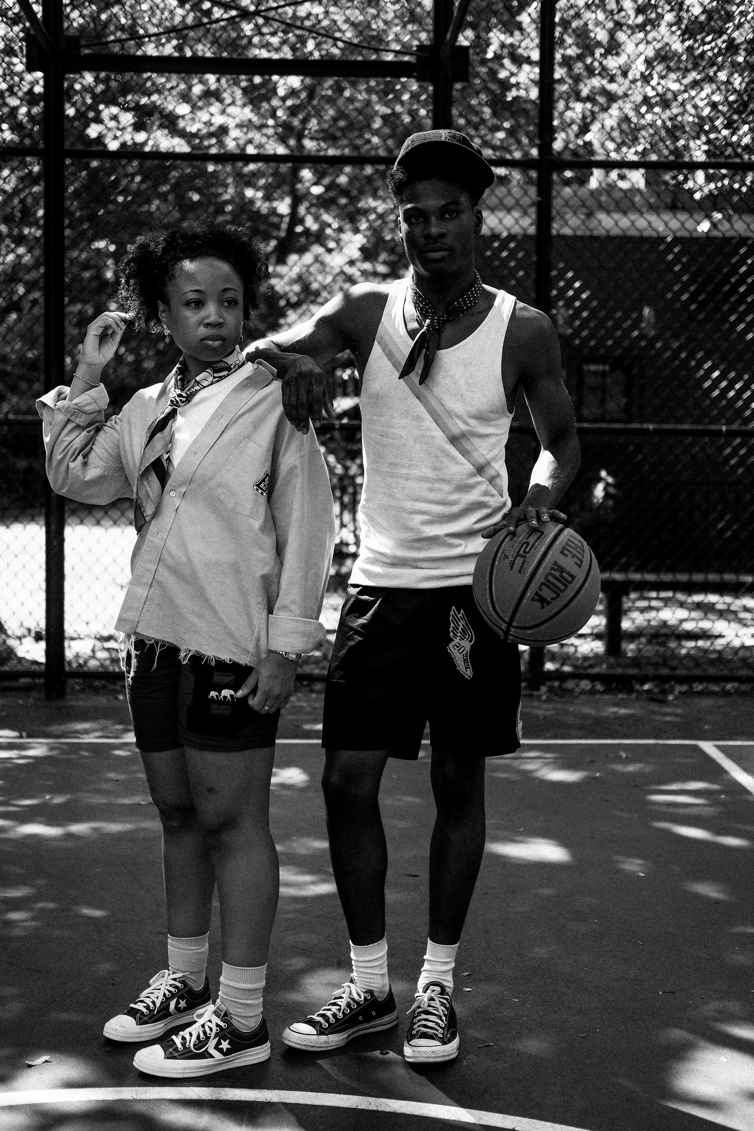 Black and white photo of a young woman and a young man standing on a basketball court outdoors. The young woman is adjusting her hair and wearing casual streetwear, and the young man is holding a basketball, wearing a tank top, shorts, and a cap. Beh