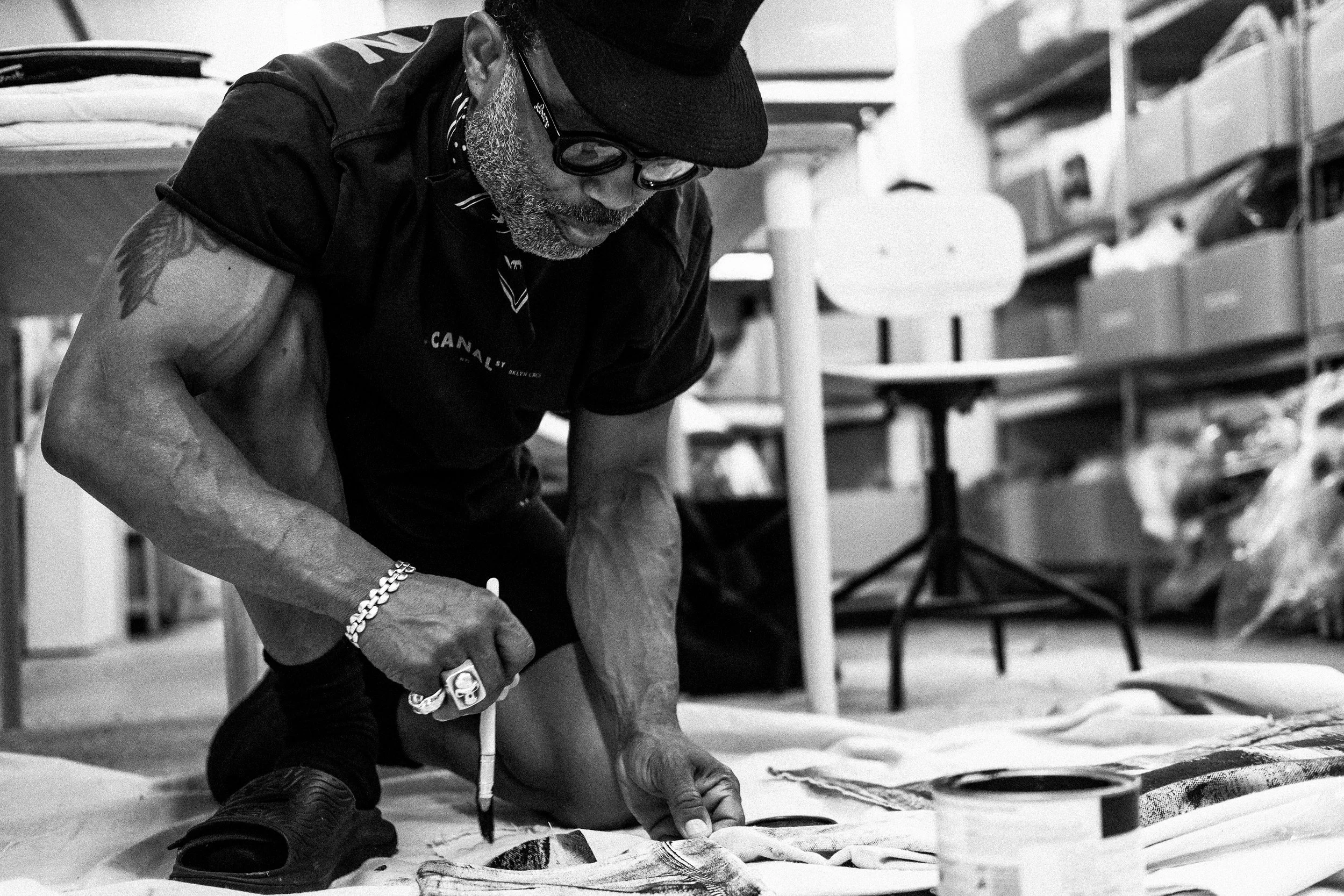 A man with tattoos, glasses, and a cap is kneeling on the floor, working on a piece of art or craft, with shelves and a lamp in the background.