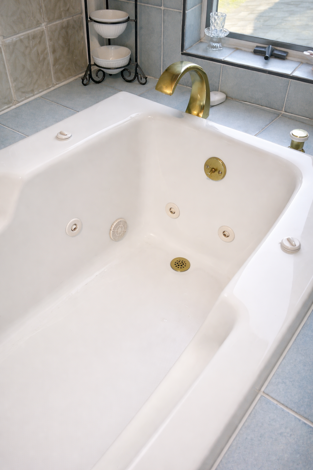 Close-up of a white bathtub with gold fixtures and jets, located in a bathroom with gray tiles and a window.