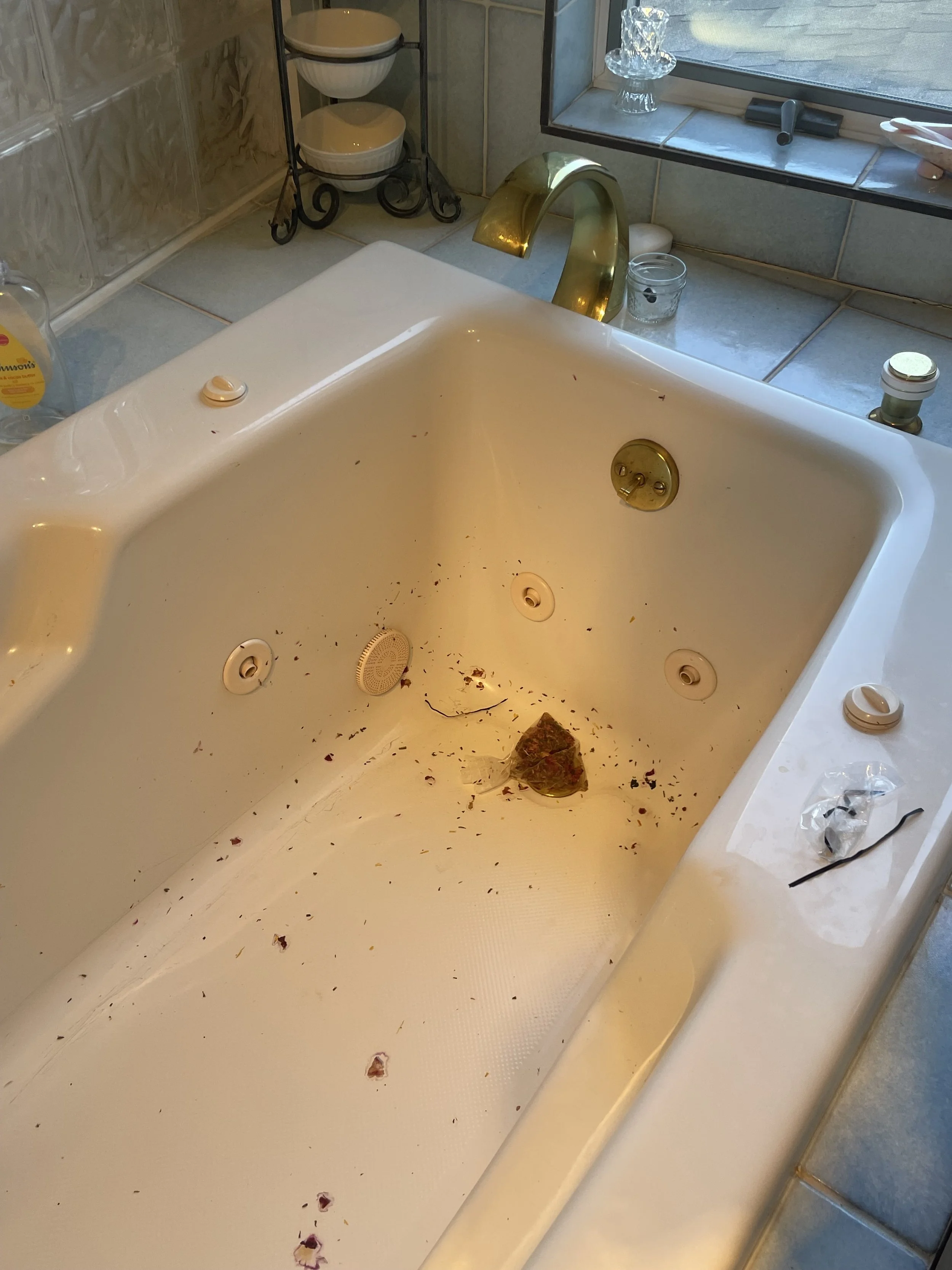 Dirty bathtub with a stone, debris, and black particles inside, surrounded by various bathroom items and a window.