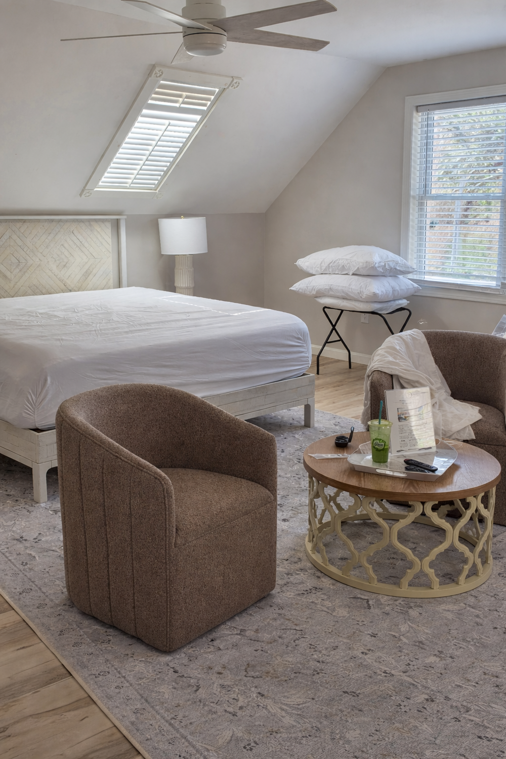 A cozy bedroom with a white bed, a table with pillows on it, a armchair, a coffee table with a drink and remote, and a window with blinds letting in natural light.