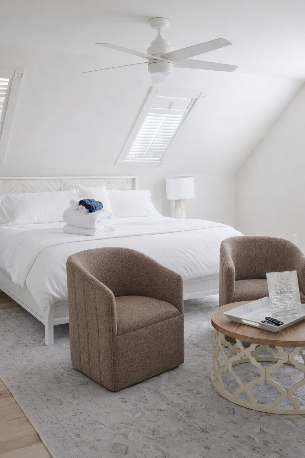 A clean, bright bedroom featuring a white bed with rolled towels on top, two brown upholstered chairs, a round wooden table, a white lamp, and a ceiling fan, with a skylight window in the sloped ceiling.