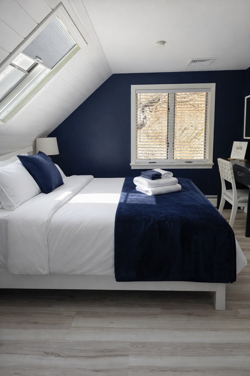 A neatly made bed with white sheets, dark blue pillows, and a dark blue blanket in a cozy bedroom with a sloped ceiling, a window with blinds, a lamp, and a desk with a chair.
