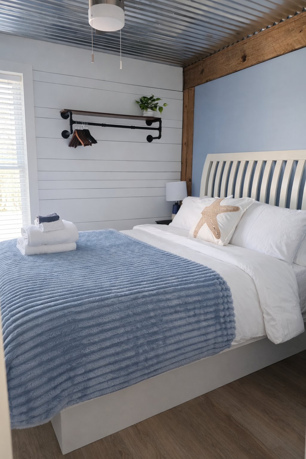 A bedroom with a white bed, light blue throw blanket, starfish pillow, and folded towels on the bed. A nightstand with a lamp is next to the bed. The wall behind the bed has a wooden beam and a blue accent wall. A black pipe clothing rack with hanger