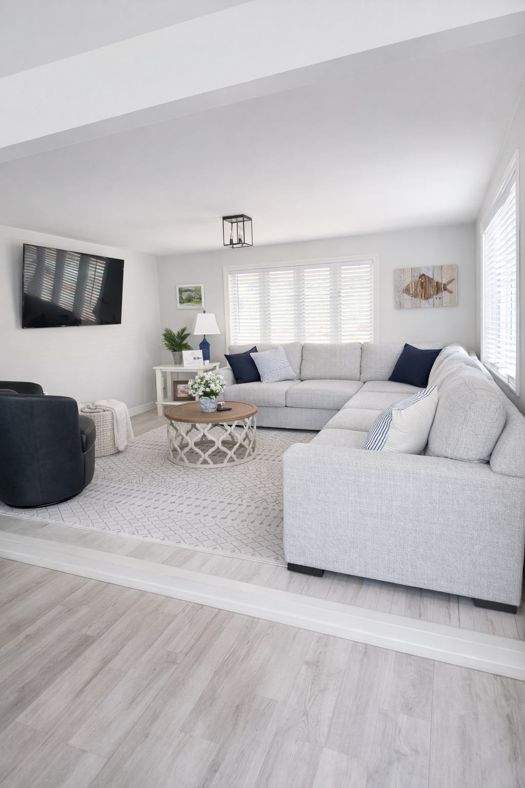 Bright living room with white walls, large window, white sectional sofa with navy and striped pillows, black armchair, round wooden coffee table with flower vase, TV mounted on wall, and decorative art on walls.