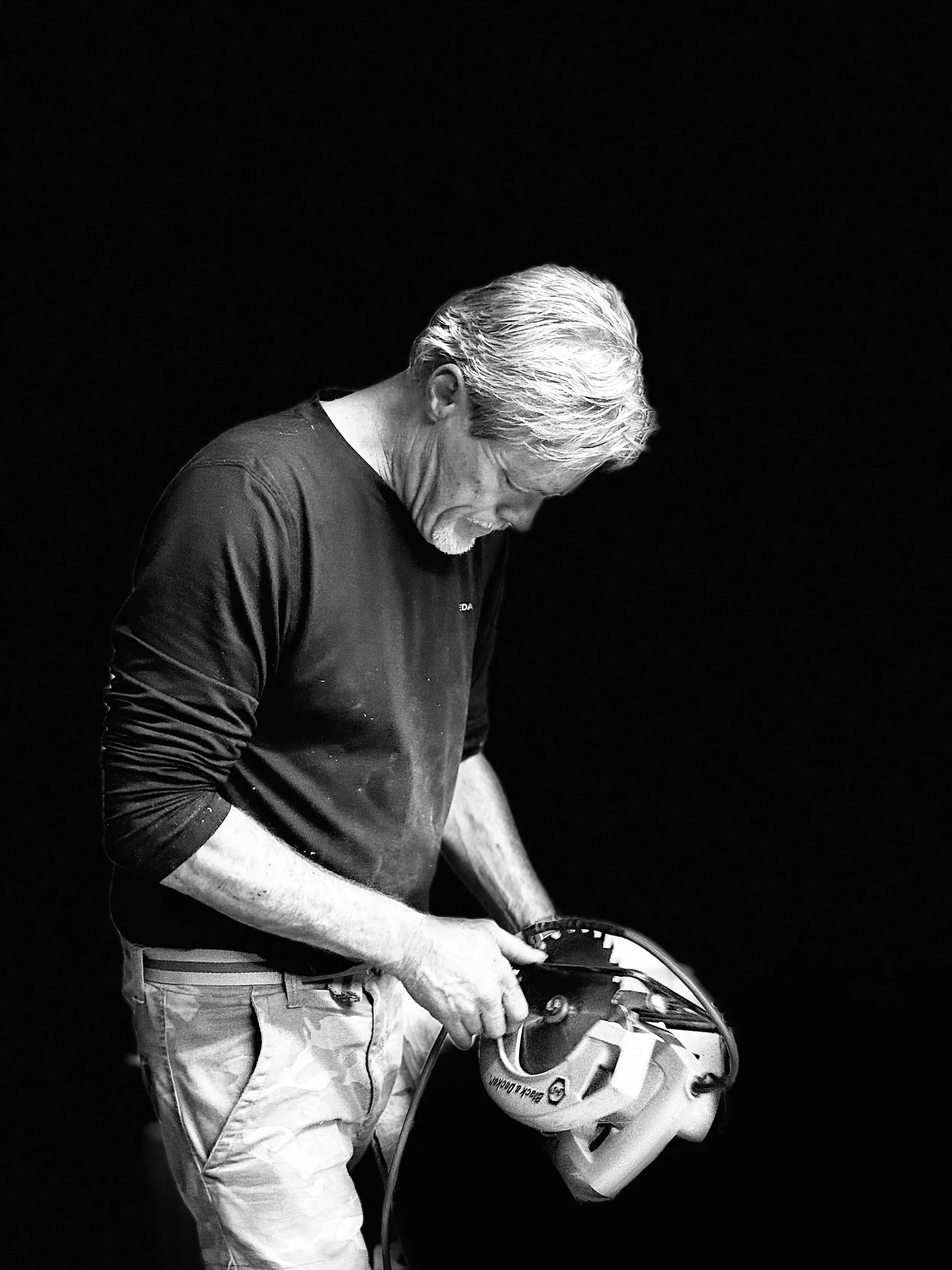 An elderly man with gray hair and a beard holding a power tool, focused on his work, dressed in a dark long-sleeve shirt and light-colored pants, against a dark background.