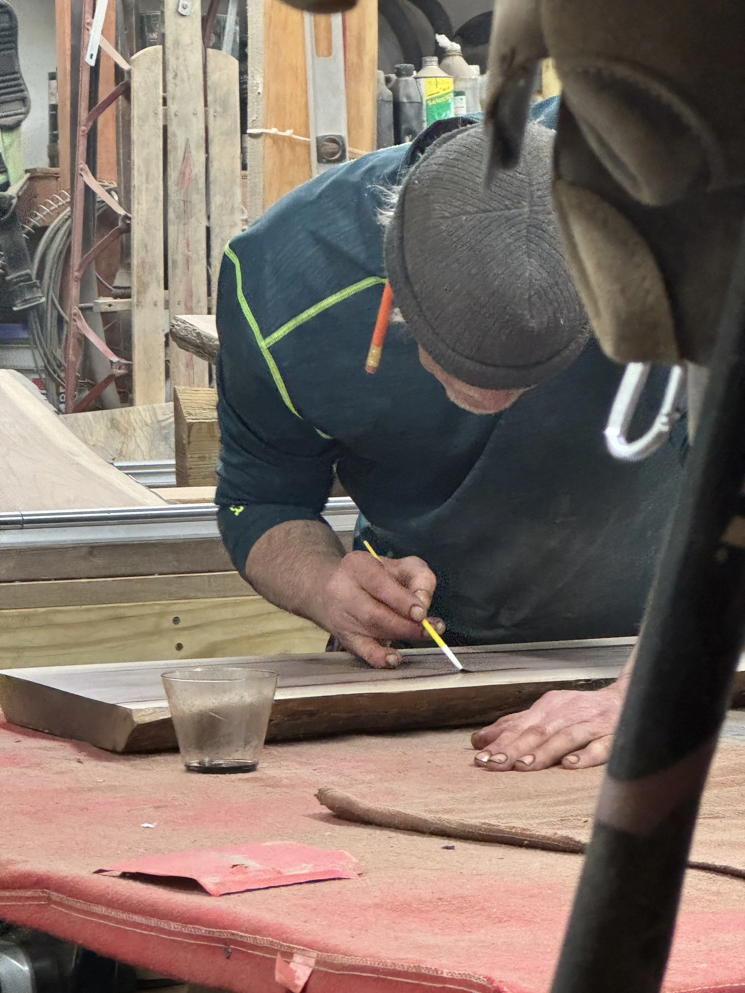 A man wearing a beanie and a dark jacket is working on a woodworking project, marking a piece of wood with a pencil in a workshop.