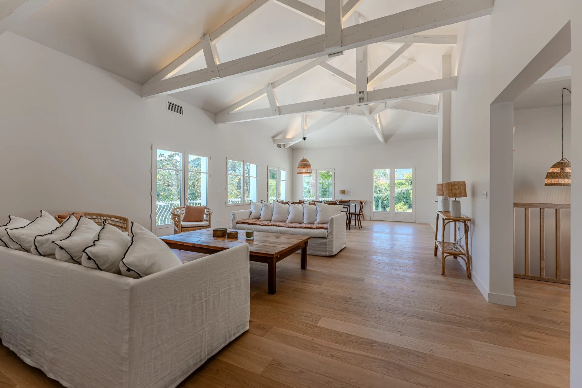 Intérieur d'un salon lumineux avec canapés blancs, tables en bois, fenêtres grandes laissant entrer la lumière naturelle, et plafond en poutres apparentes blanches.