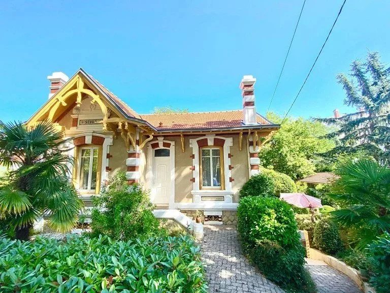 Une maison ancienne avec une façade en pierre verte, entourée de végétation luxuriante, sous un ciel bleu clair.