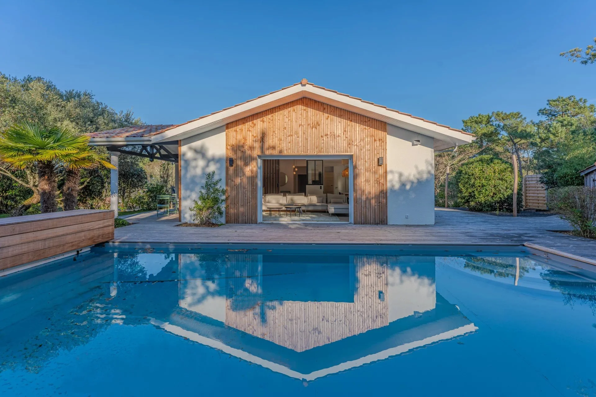 Maison moderne avec façade en bois, grande baie vitrée ouverte sur salon intérieur, piscine en premier plan, entourée de végétation et un ciel bleu clair.