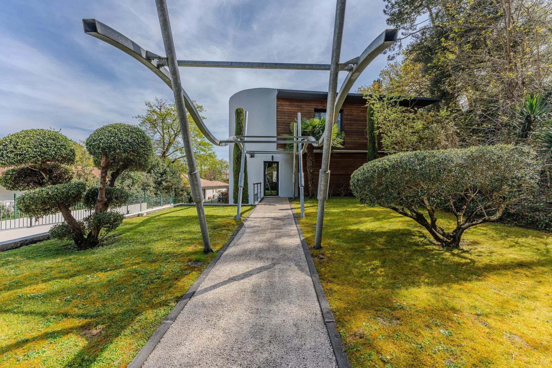 Chemin menant à une maison moderne en pierre blanche, entourée de jardin avec arbres taillés et herbe verte, ciel partiellement nuageux.