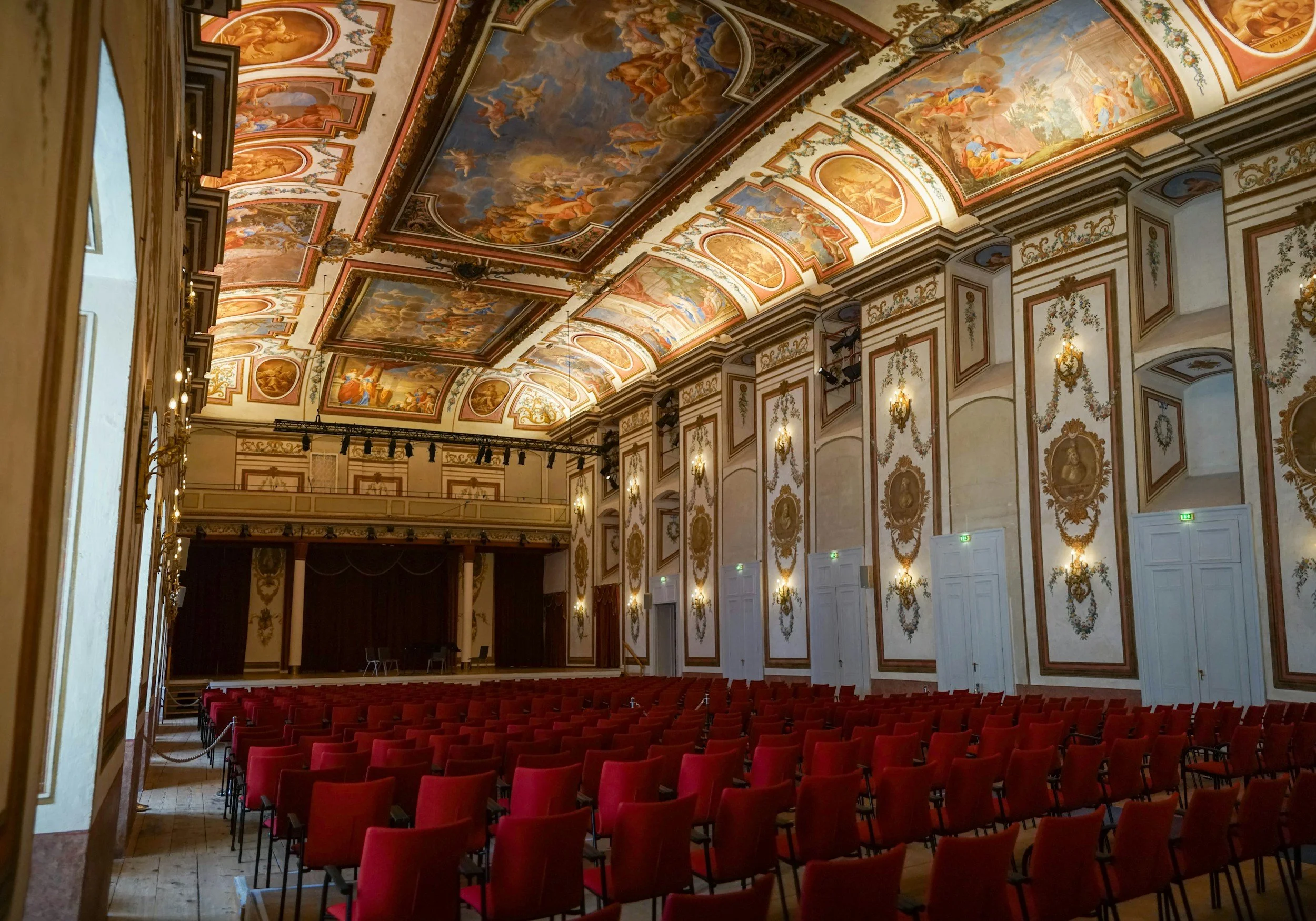 Salle de concert ornée de fresques artistiques au plafond, avec des murs décorés de motifs dorés et de portraits en ovales, chaises rouges alignées face à une scène vide.