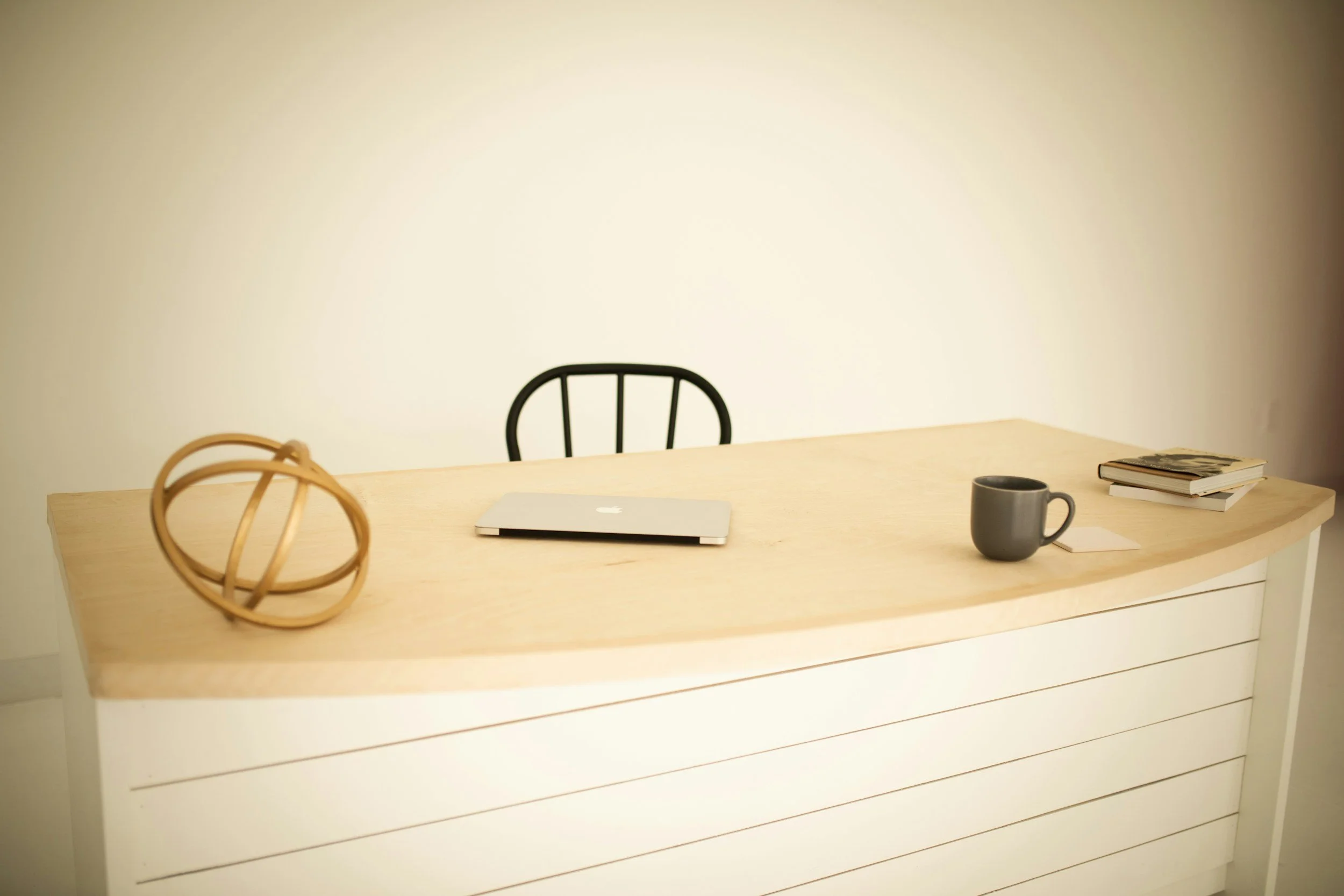 Bureau minimaliste avec une table en bois clair, une ordinateur portable, une tasse grise, une pile de livres, une décoration en forme de sphère en bois, et une chaise noire en arrière-plan.