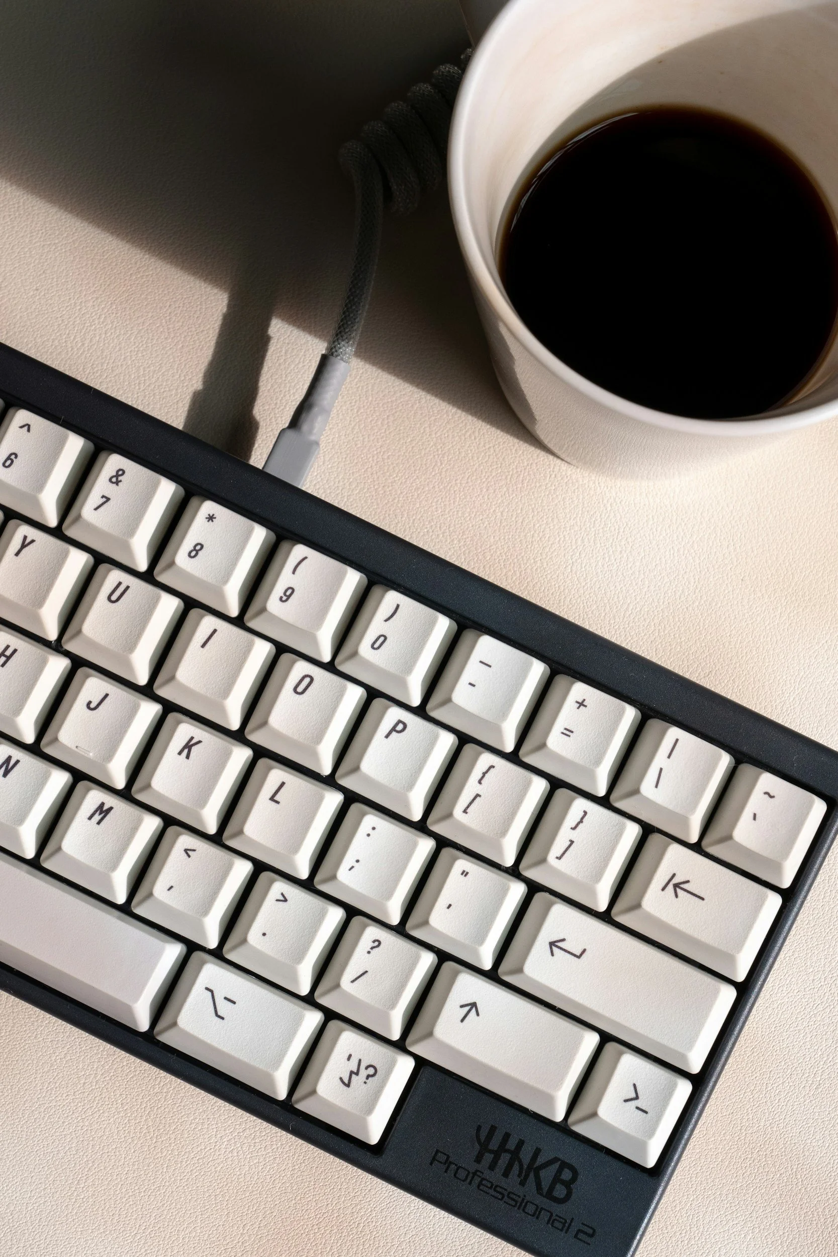 Clavier d'ordinateur blanc avec marque YHKB, un câble connecté, et une tasse de café noir sur une surface beige.