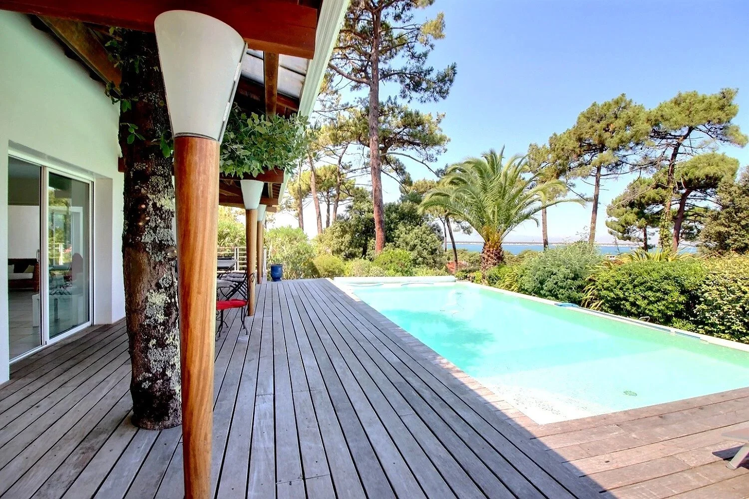 Terrasse en bois avec piscine, entourée d'arbres et de végétation, vue sur l'océan en arrière-plan, partie d'une maison moderne avec une grande fenêtre.