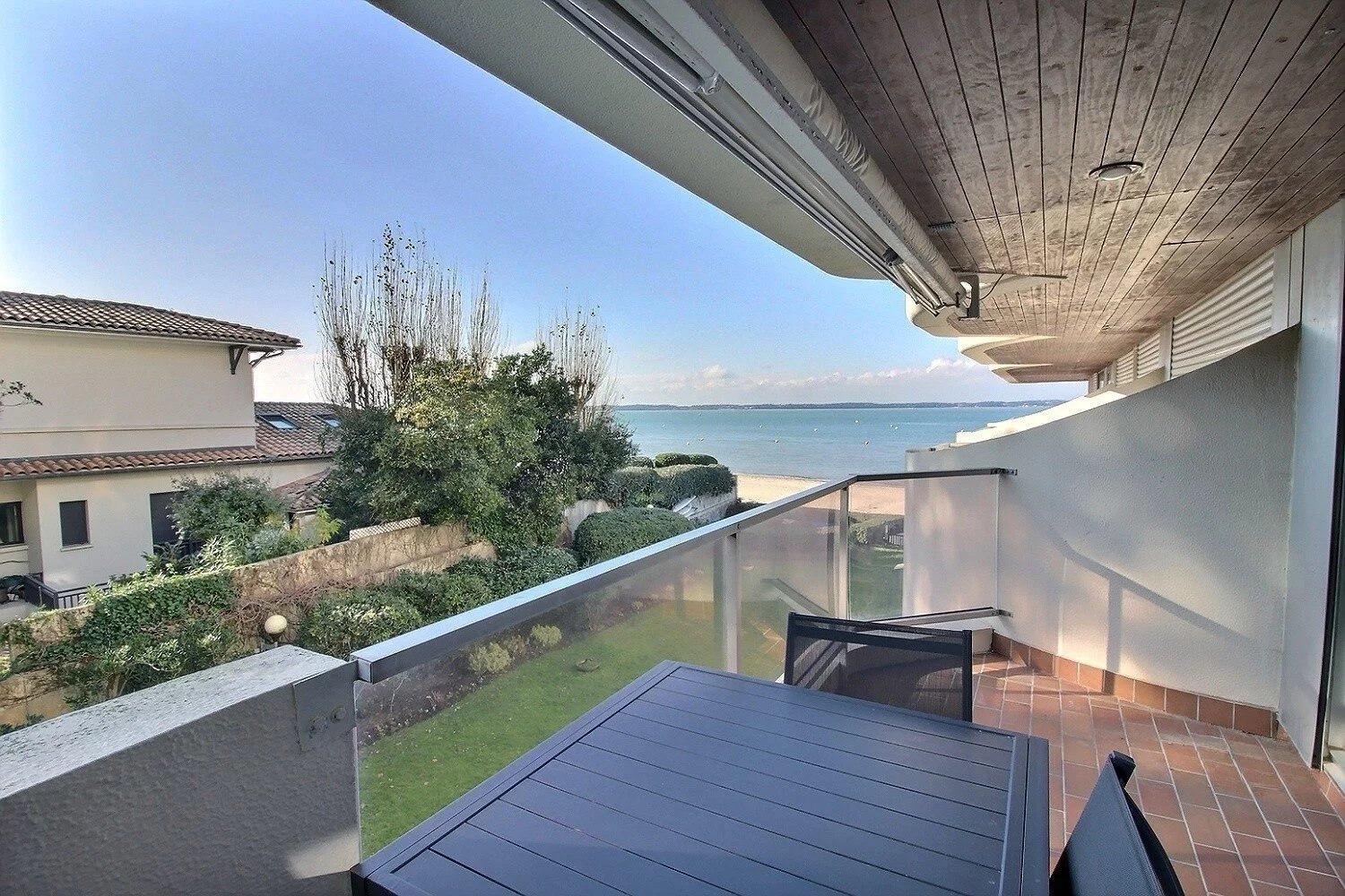 Balcon avec table et chaises, vue sur la mer, maisons voisines et jardins, ciel bleu et horizon éloigné.