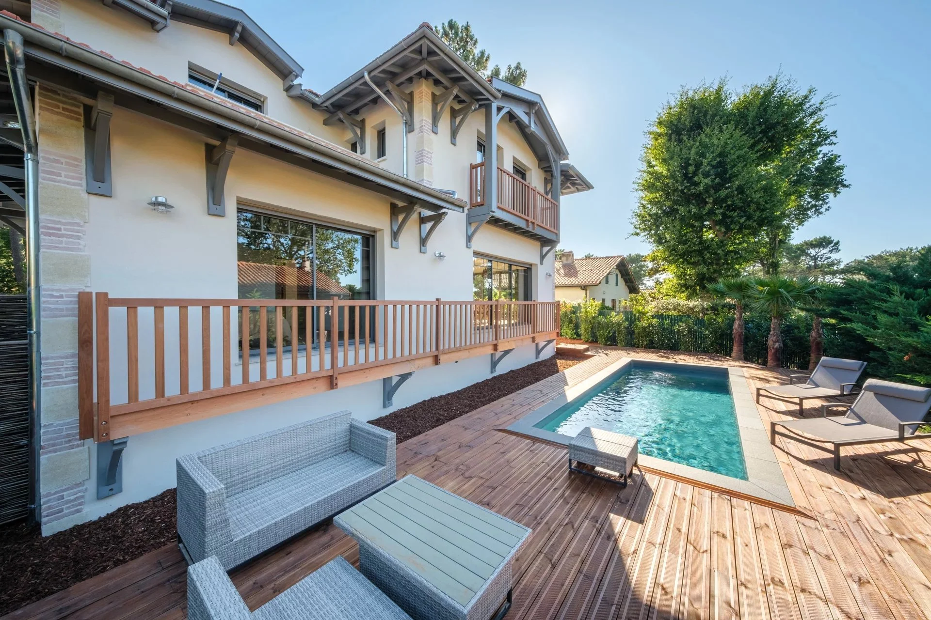 Vue d'une piscine dans la cour d'une maison avec terrasse en bois, chaises longues, et arbre verdoyant en arrière-plan, maison moderne à deux étages avec balcon en bois.