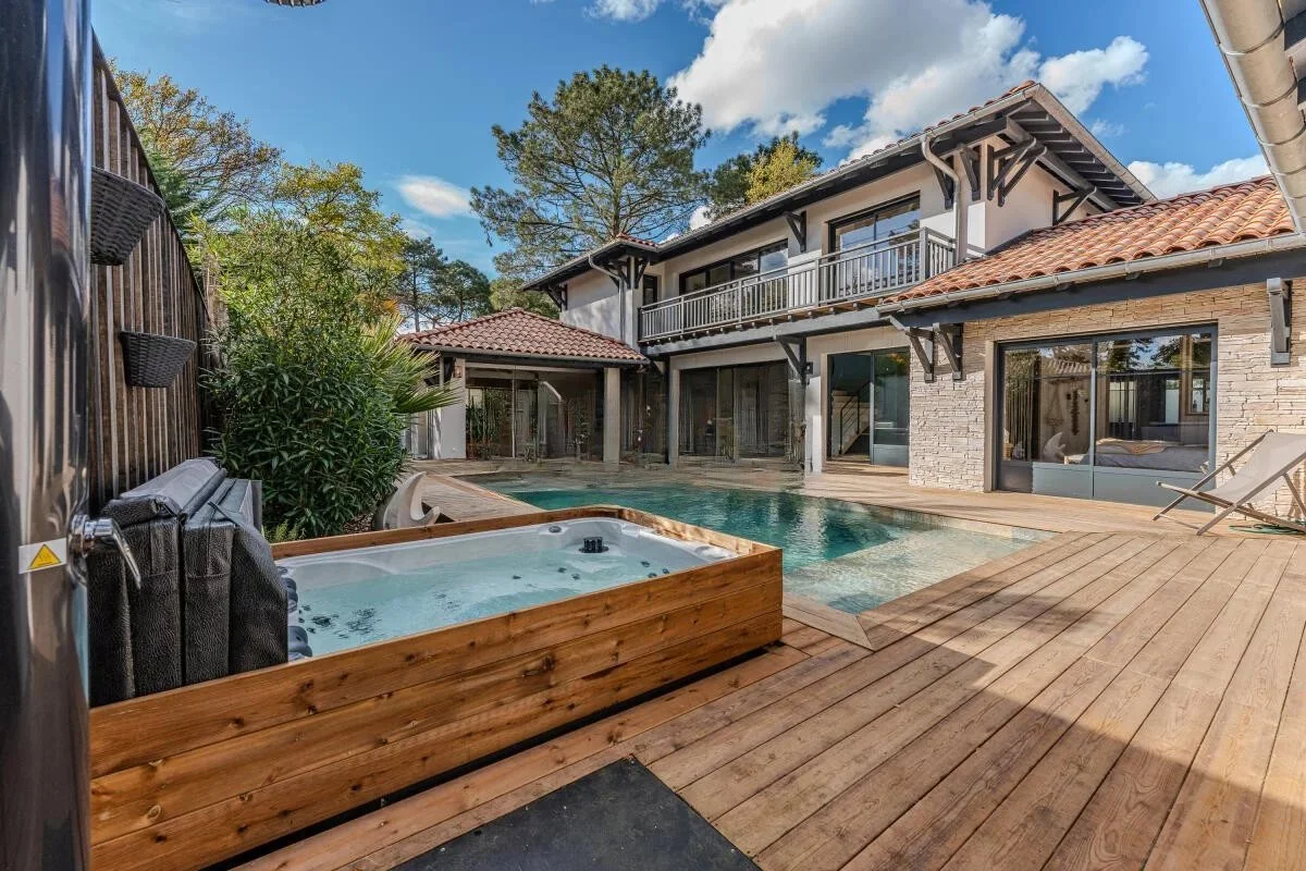Piscine extérieure avec jacuzzi en bois au premier plan, maison moderne avec deux étages et balcon en arrière-plan, entourée d'arbres, ciel bleu avec quelques nuages.