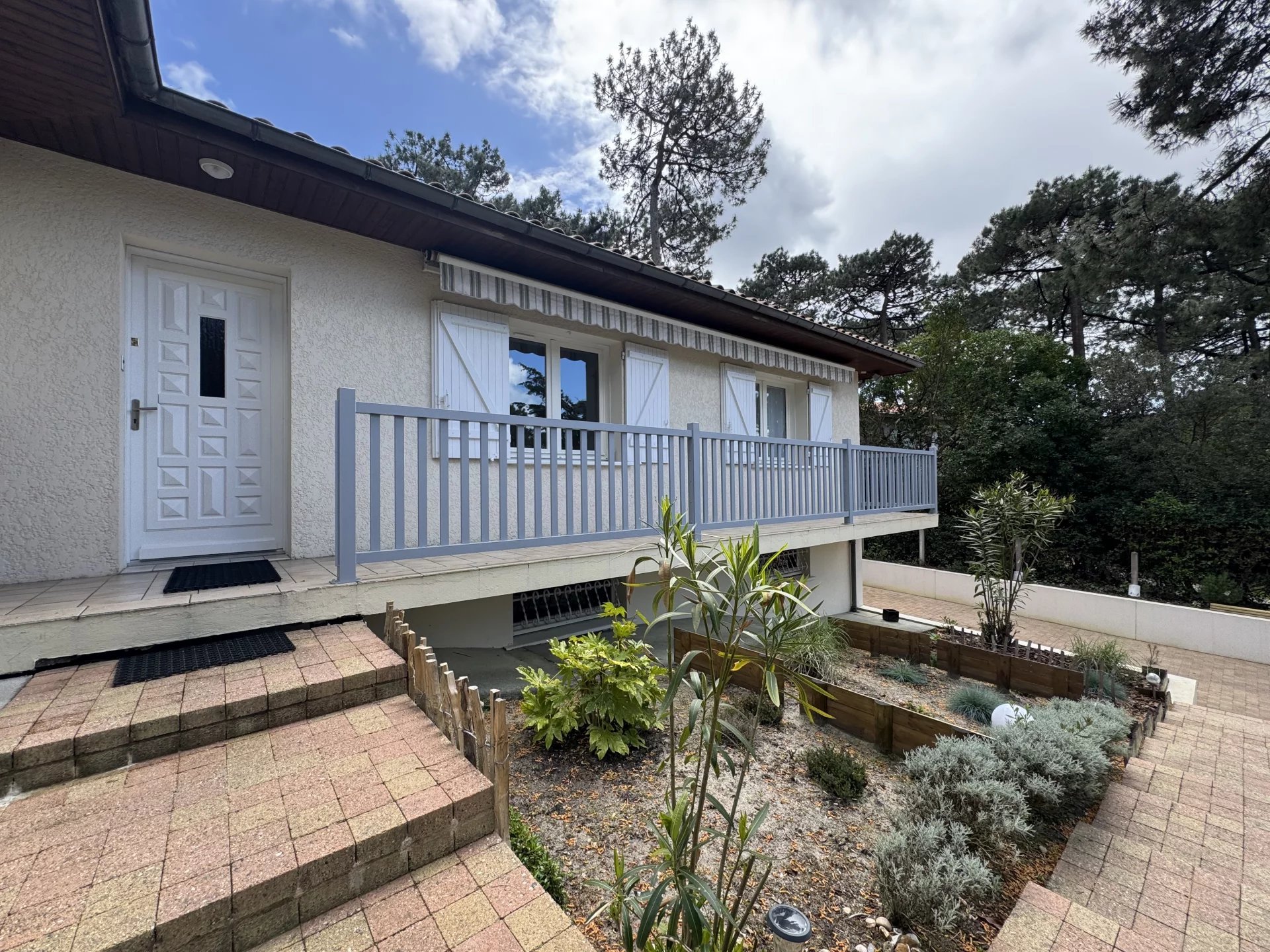 Maison avec terrasse et escalier en pierre, jardin avec diverses plantes et arbres, environnement boisé, ciel partiellement nuageux.