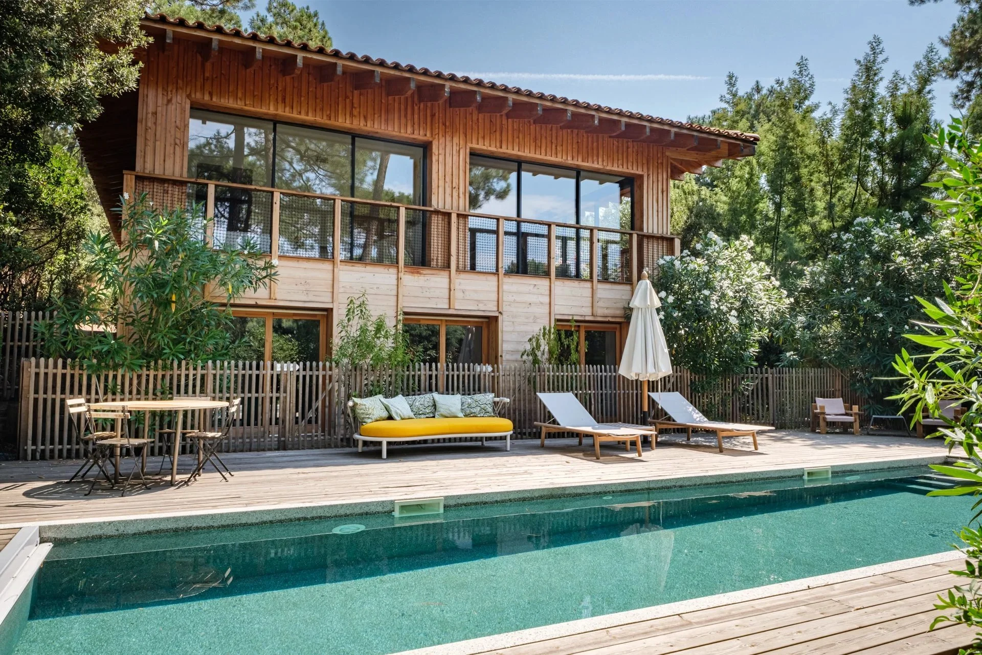 Maison en bois avec terrasse et piscine, mobilies extérieures, arbres et végétation dense, ciel bleu, ambiance relaxante