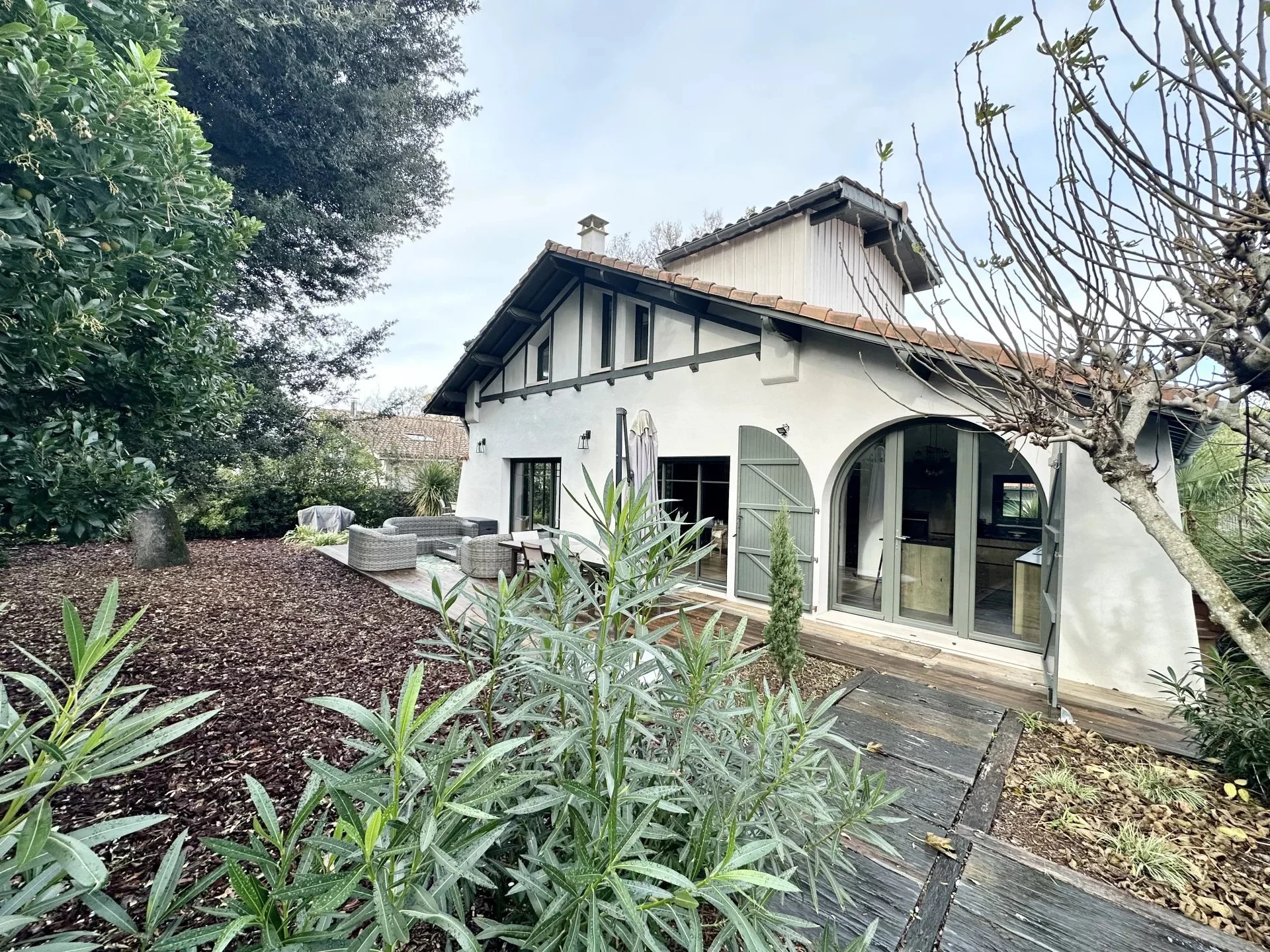 Maison à deux étages avec mur blanc, fenêtres à volets gris, porte en arc, terrasse avec mobilier extérieur, entourée de jardin avec plantes et arbres.